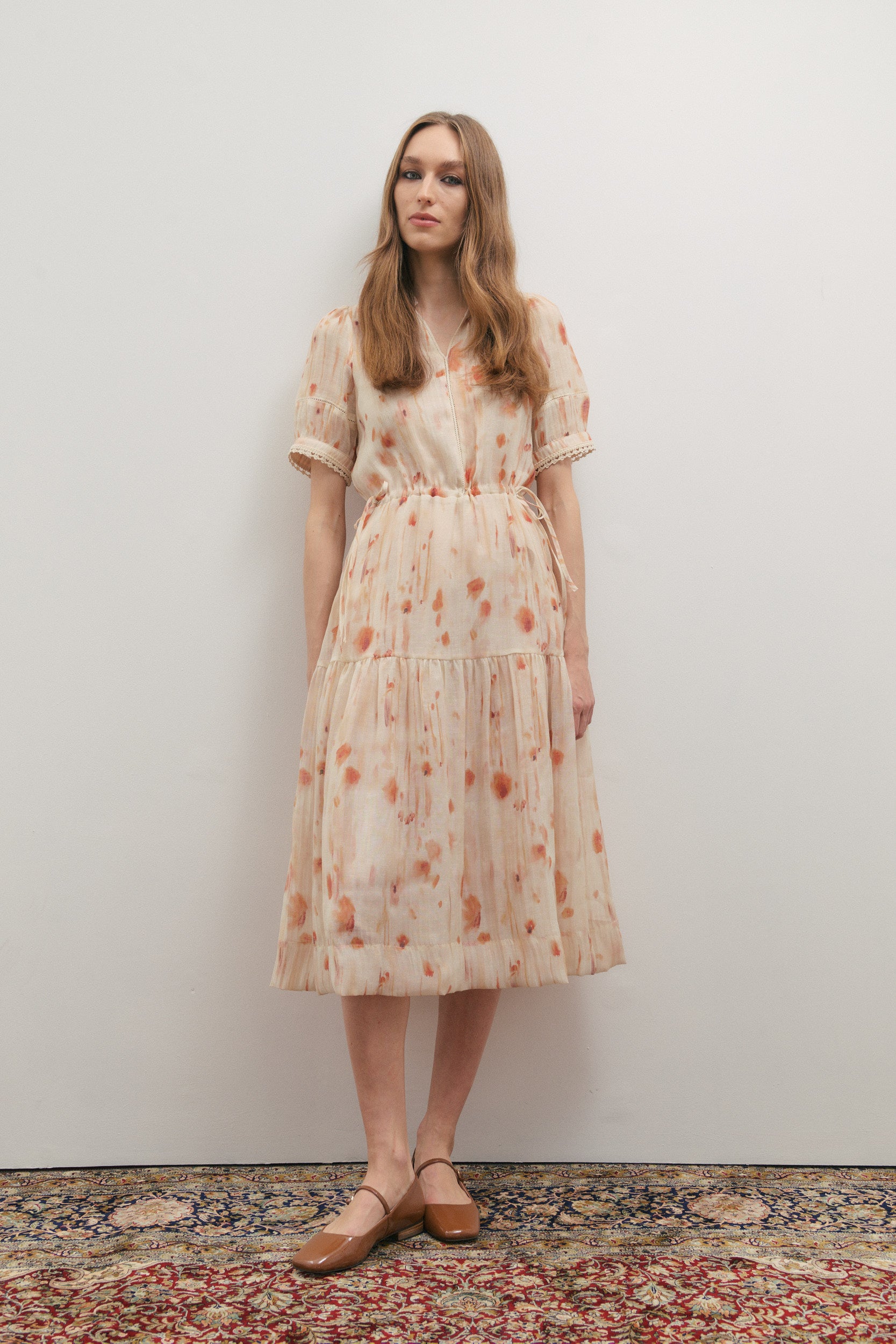 Woman wearing a floral dress standing against a plain wall with a patterned rug on the floor.
