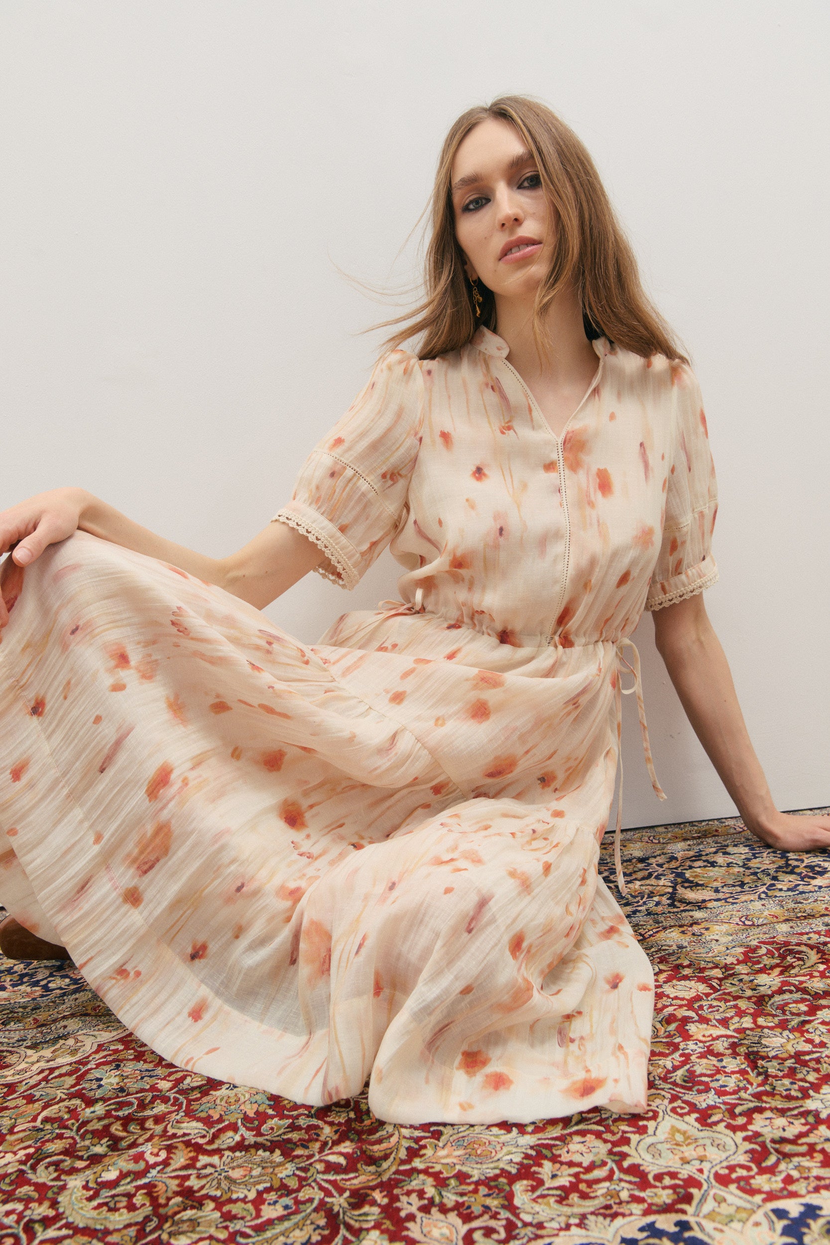 Woman in a floral dress sitting on a patterned rug against a white wall