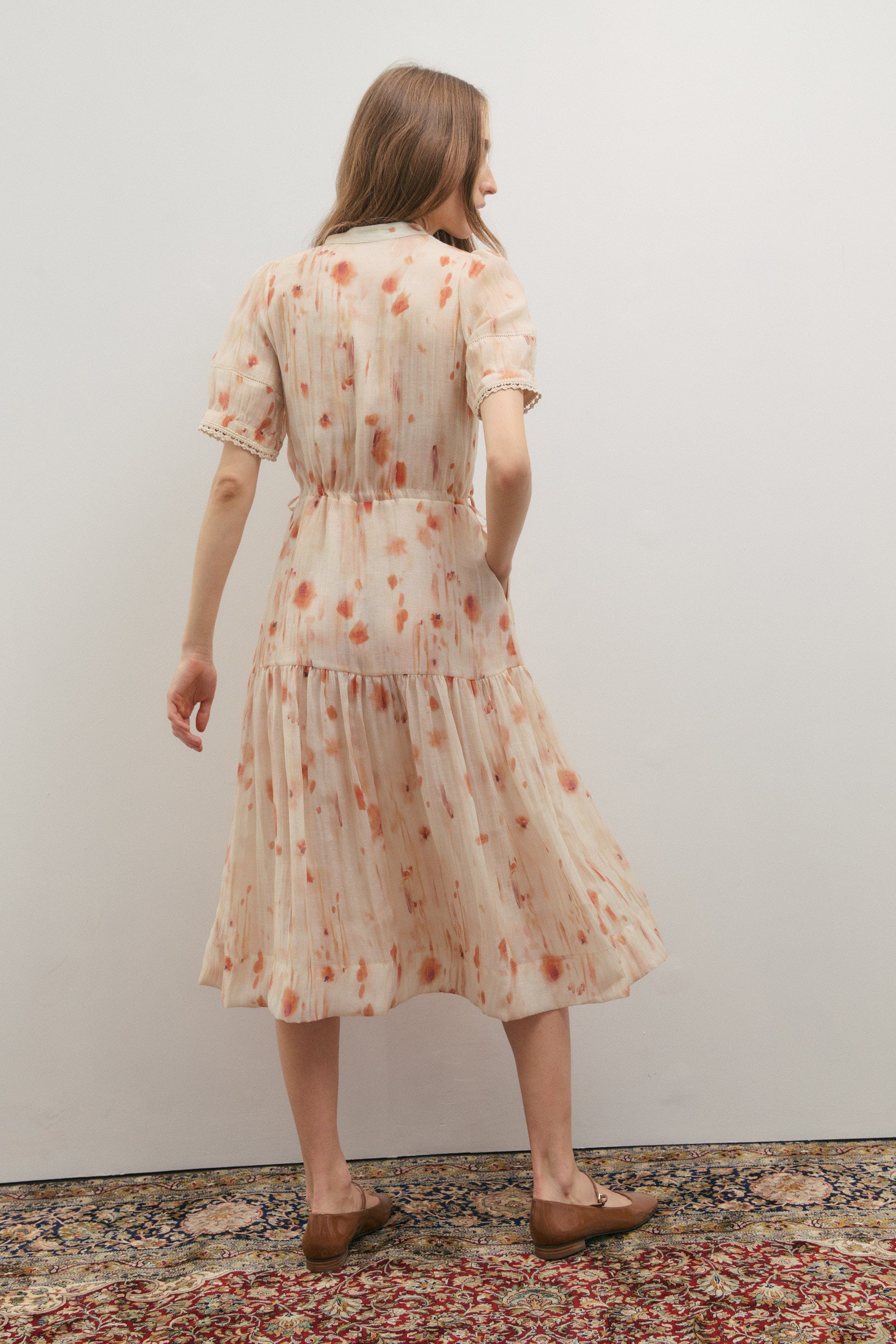 Woman wearing a floral dress standing on a patterned rug with a plain wall background