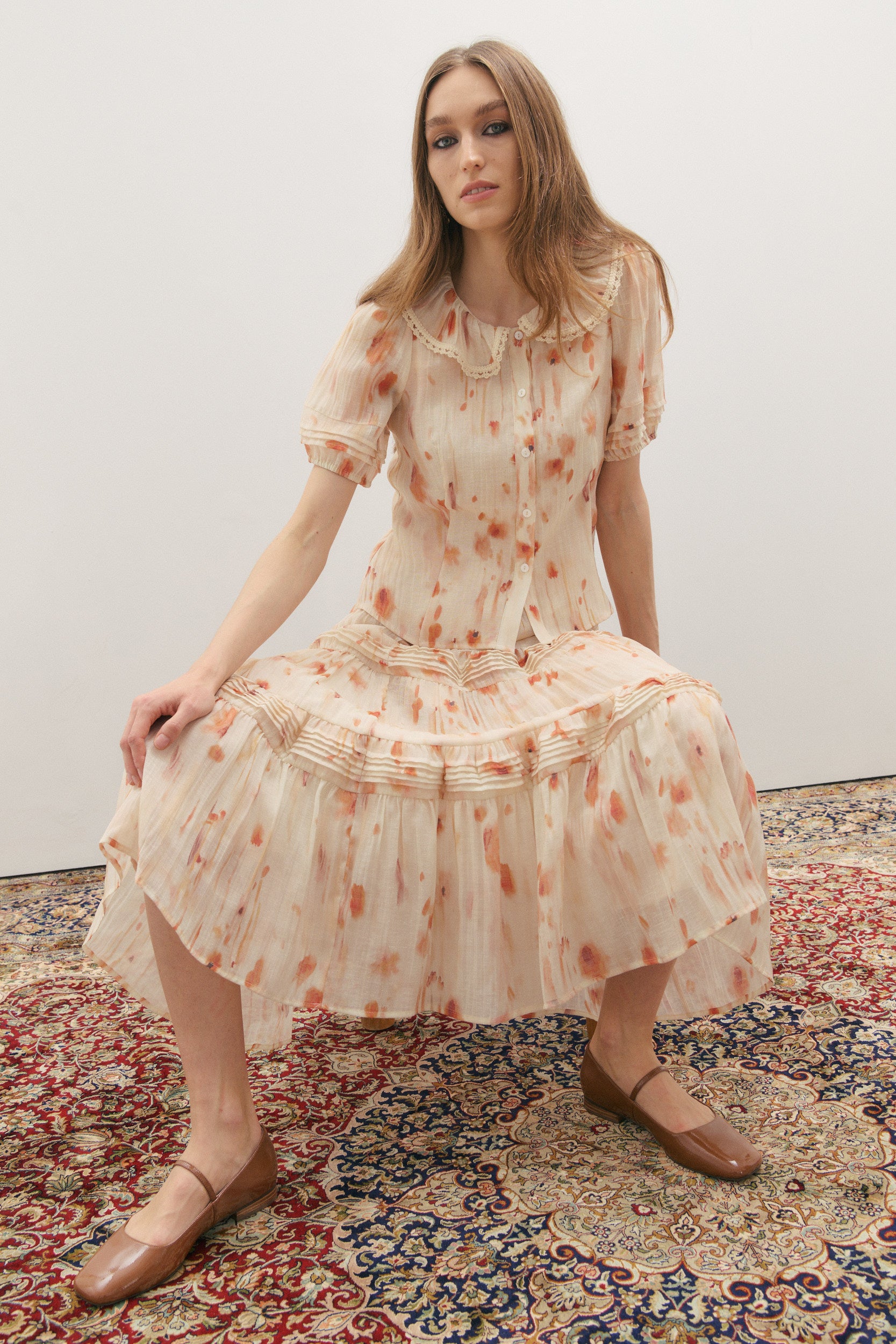 Woman wearing a cream and peach watercolour floral print skirt with pleated rows and a matching short sleeve shirt with a lace trim round collar and pleated sleeve sitting on a patterned rug.