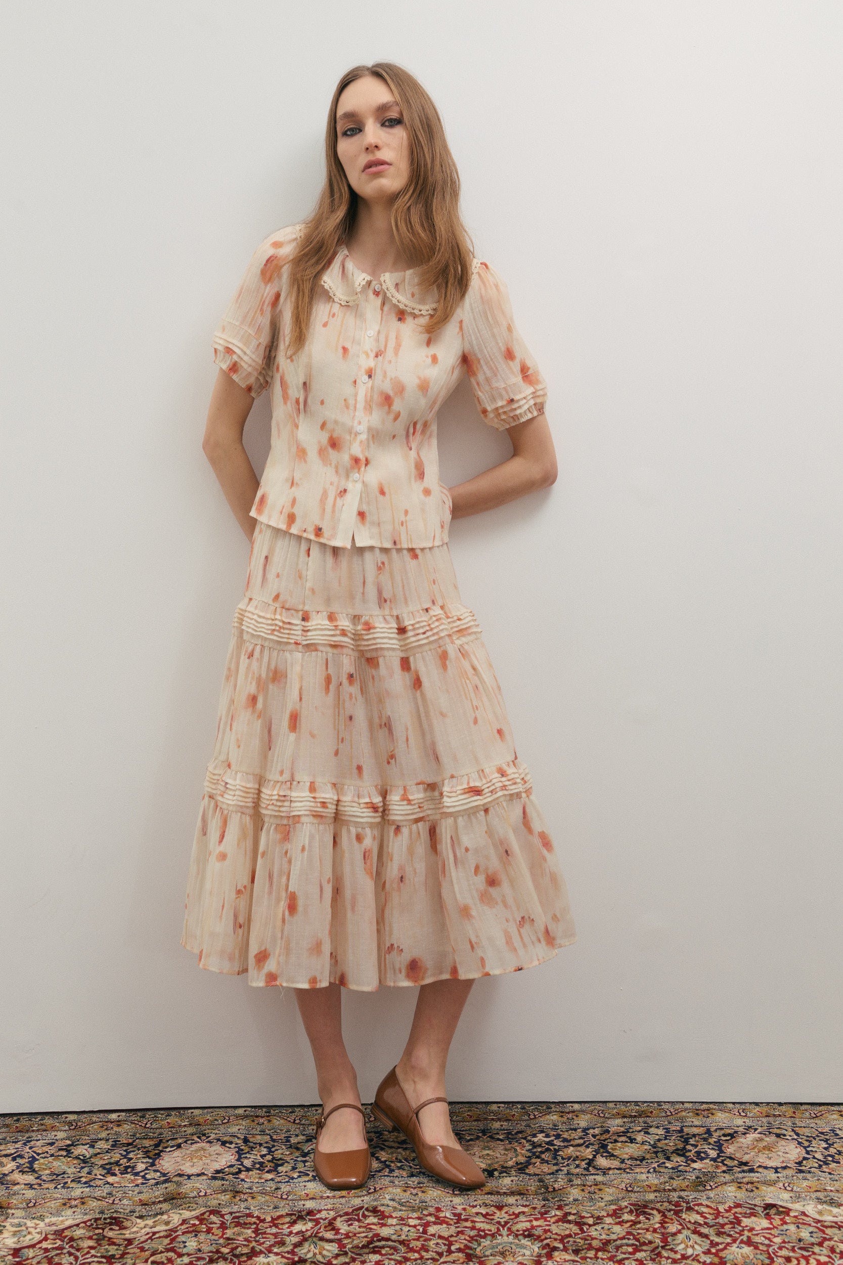 Woman wearing a floral skirt and shirt standing against a plain wall.