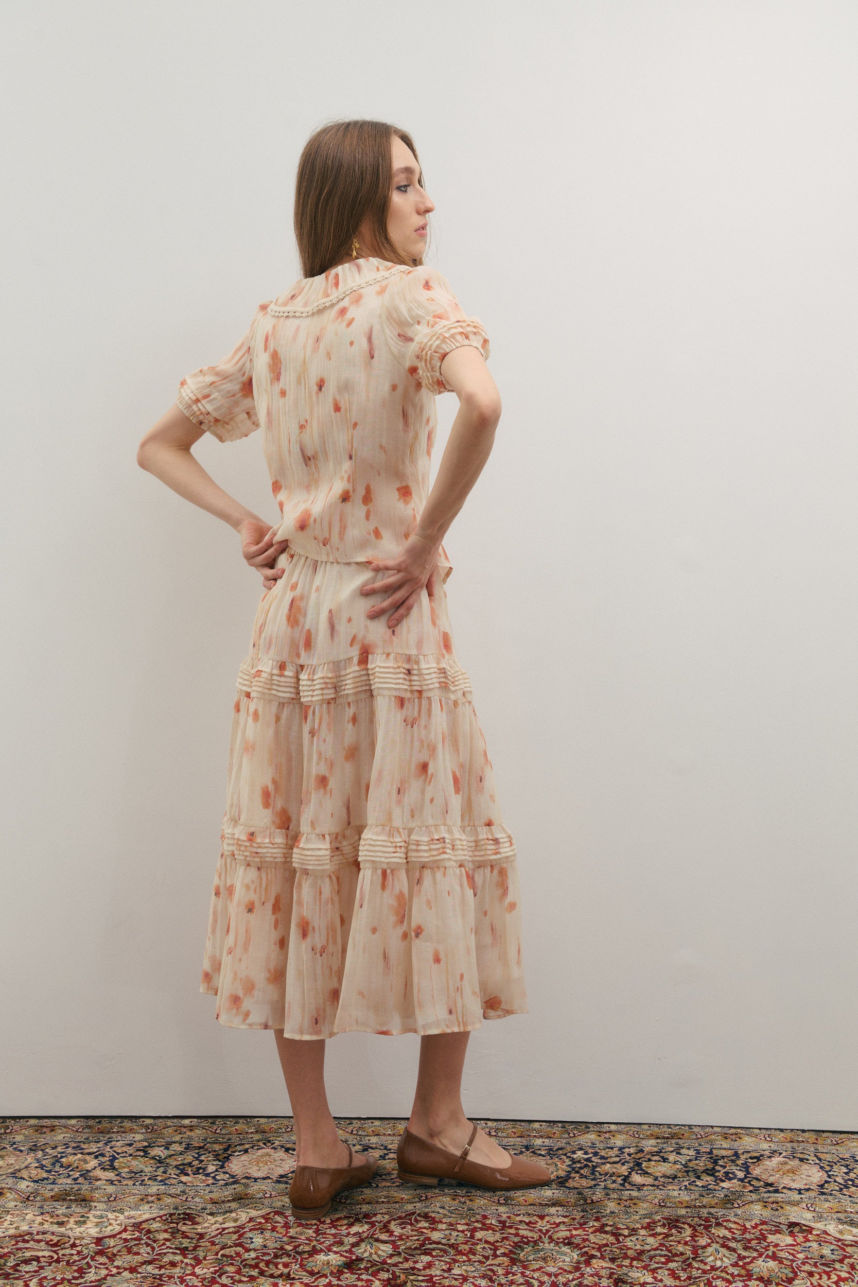 Woman wearing a floral midi skirt with rows of pleated volume with a matching short sleeve shirt by Australian fashion brand Maevarii standing against a plain wall.