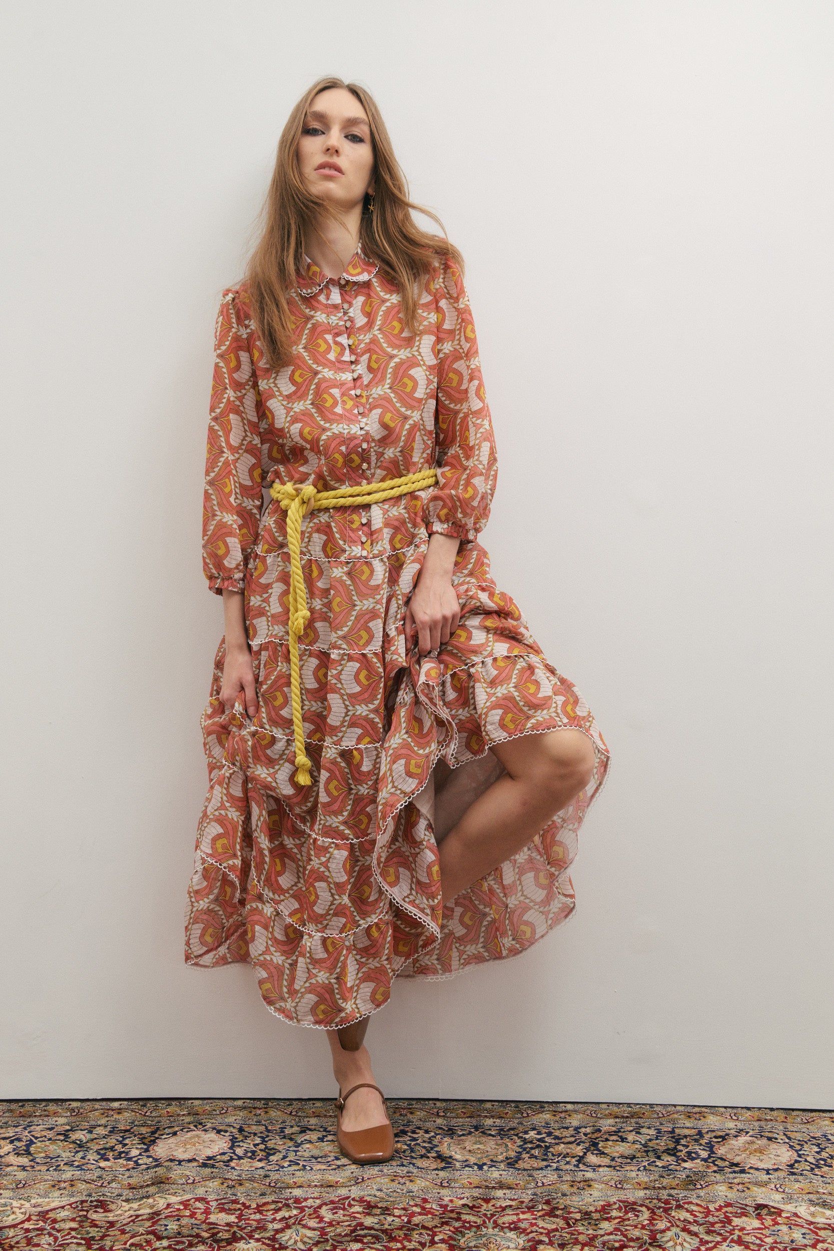 Woman wearing a patterned dress with a yellow belt against a white wall.