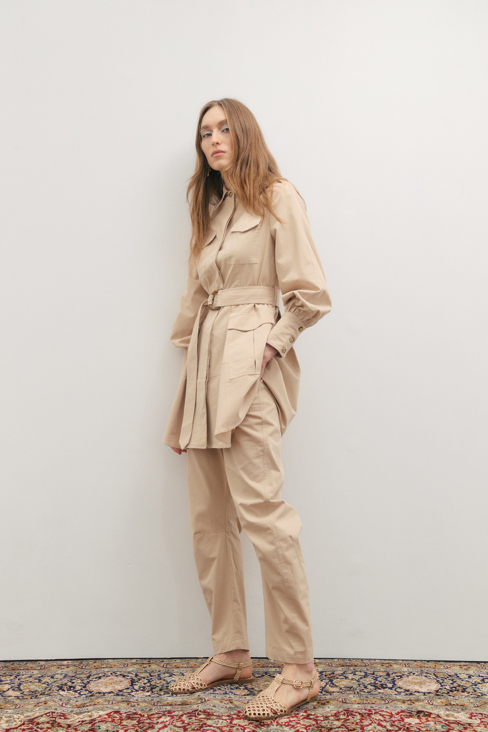 Woman wearing a beige trench coat and pants against a white wall.