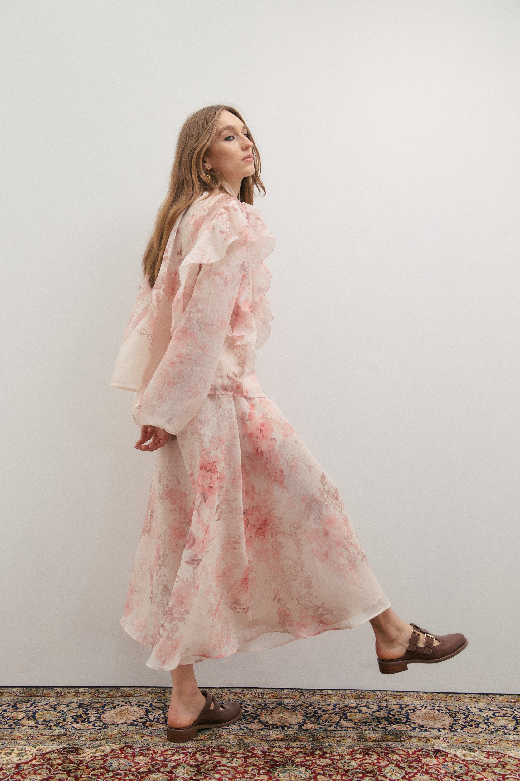 Woman wearing a pink floral maxi skirt and a matching evening jacket with long billowing sleeves and large airy ruffles with a matching neck scarf and gold and pearl button up front, standing against a white wall.