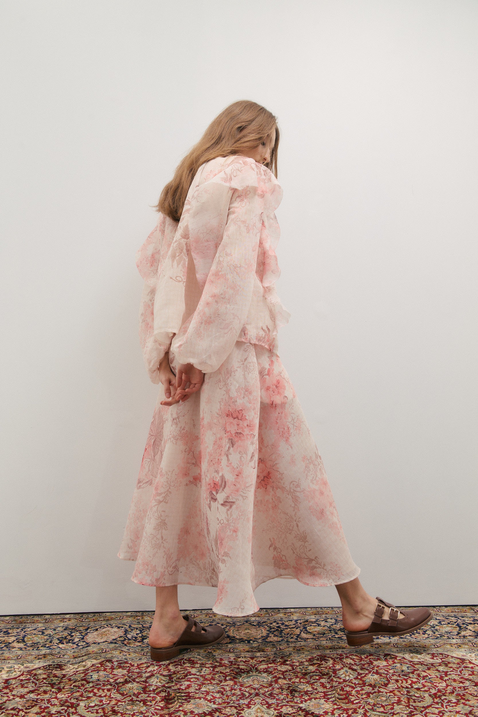 Woman wearing a floral long skirt and matching blouse standing on a patterned rug against a white wall.
