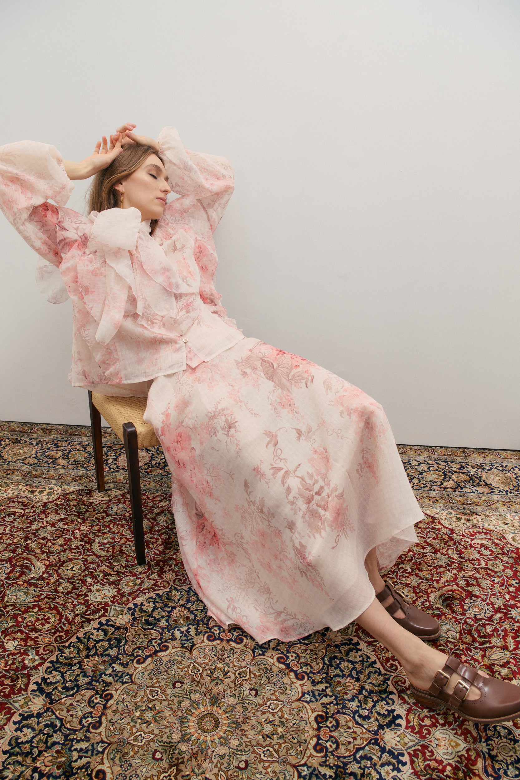 Woman in a pink floral outfit sitting on a chair with a patterned rug underneath.