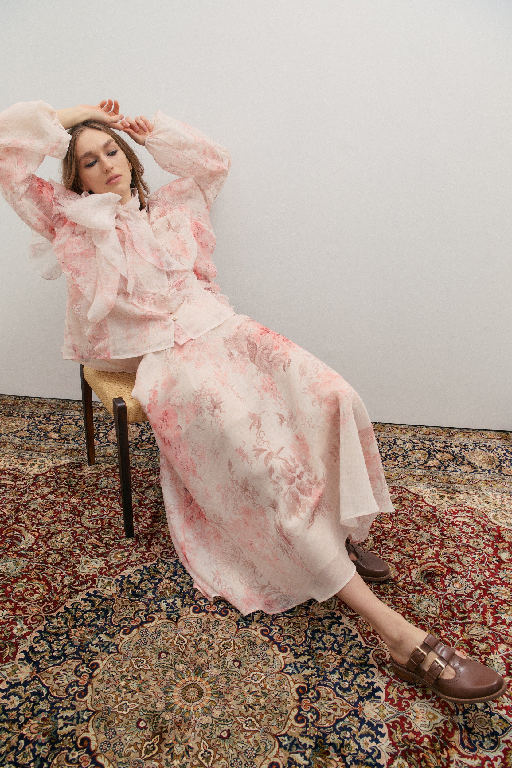 Woman in a pink floral outfit sitting on a chair with a patterned rug underneath.