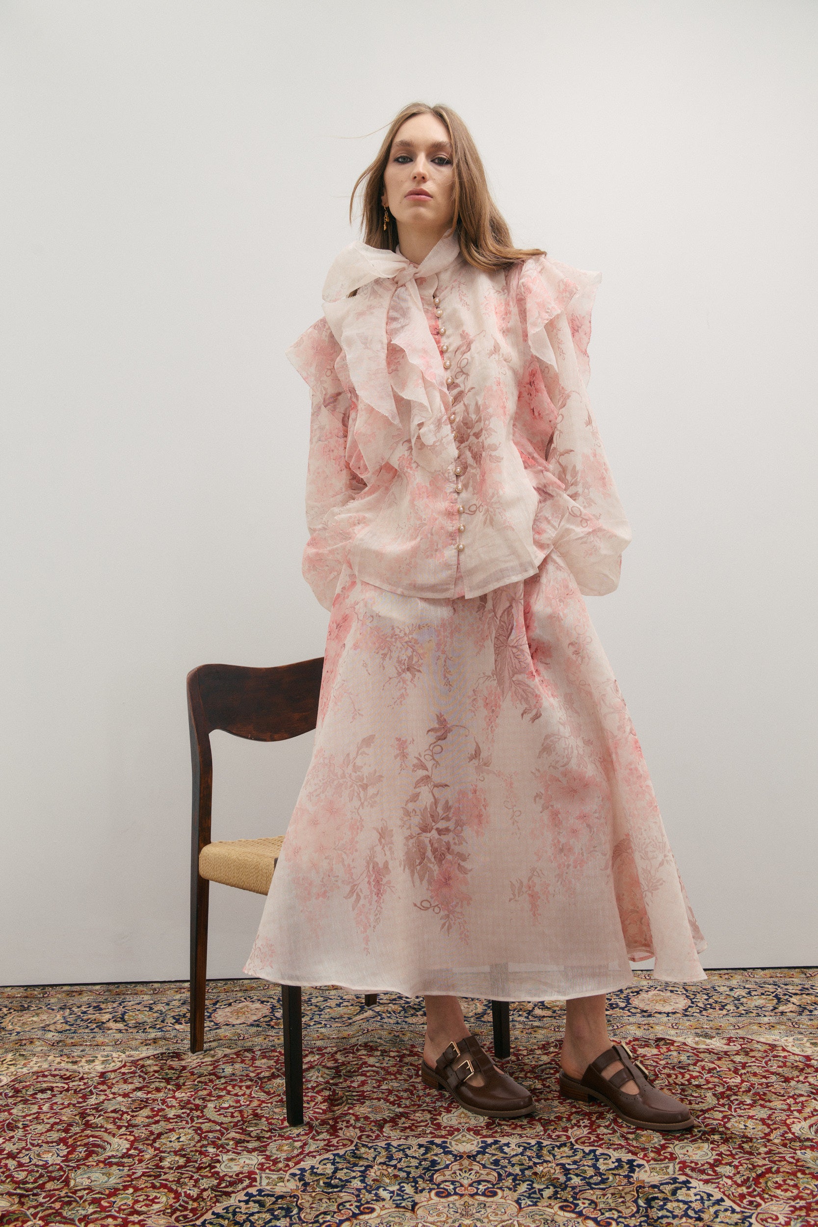 Woman in a floral skirt and frilled blouson sleeve top with neck scarf sitting on a chair against a white wall.