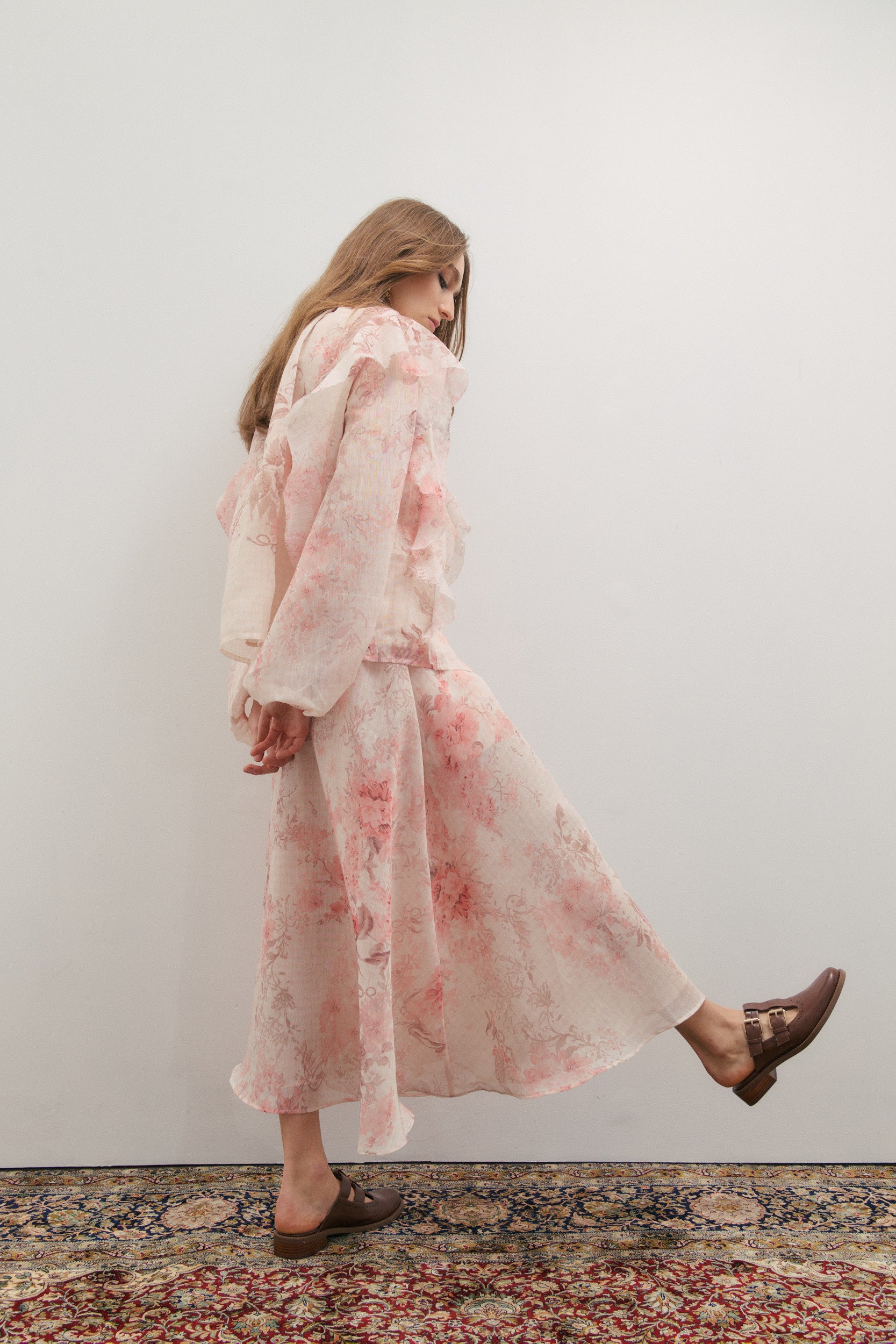Woman wearing a pink floral maxi skirt and blouse standing against a white wall.