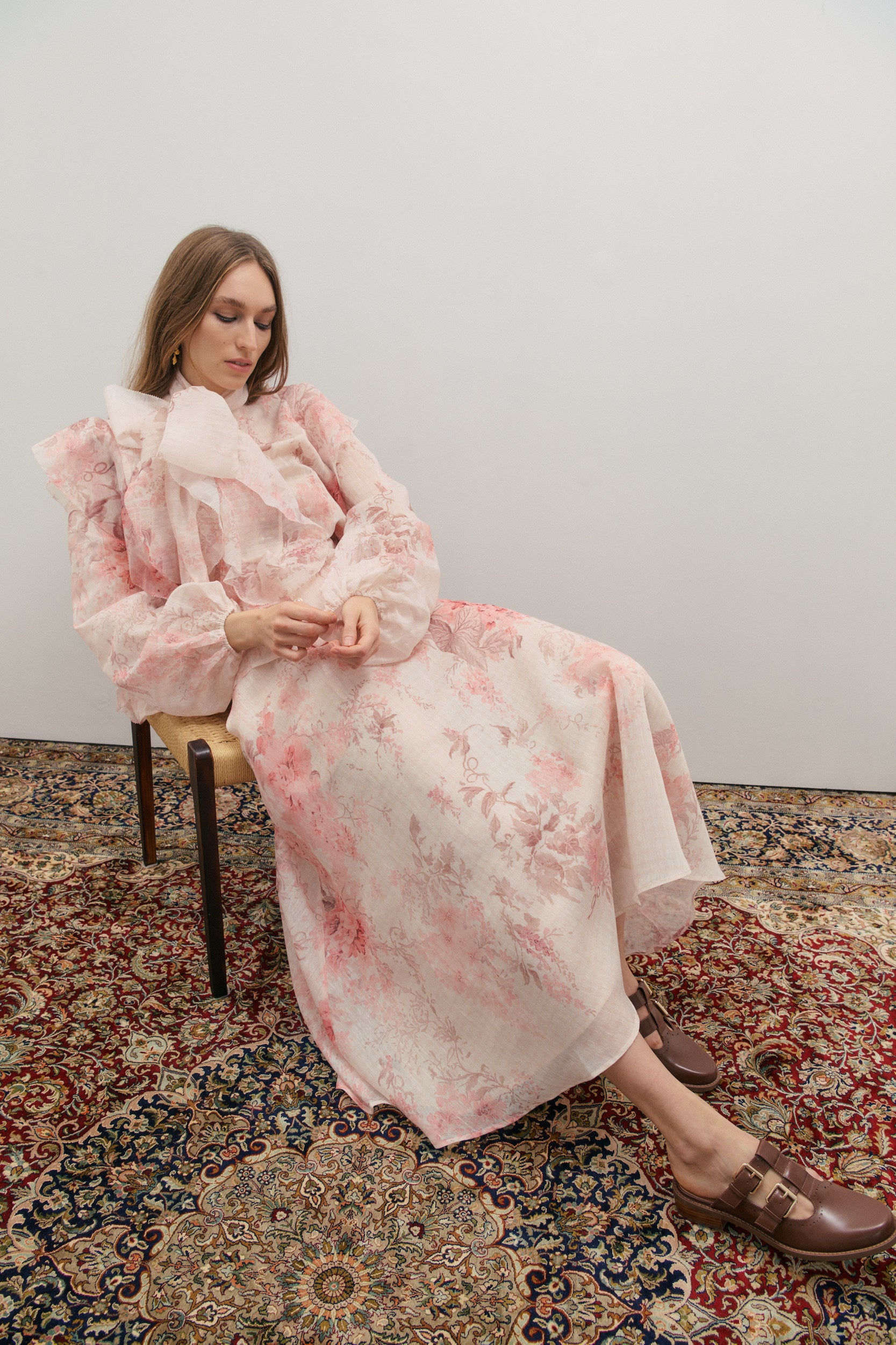 Woman in a floral skirt and blouse with matching neck scarf sitting on a chair with a patterned rug underneath.