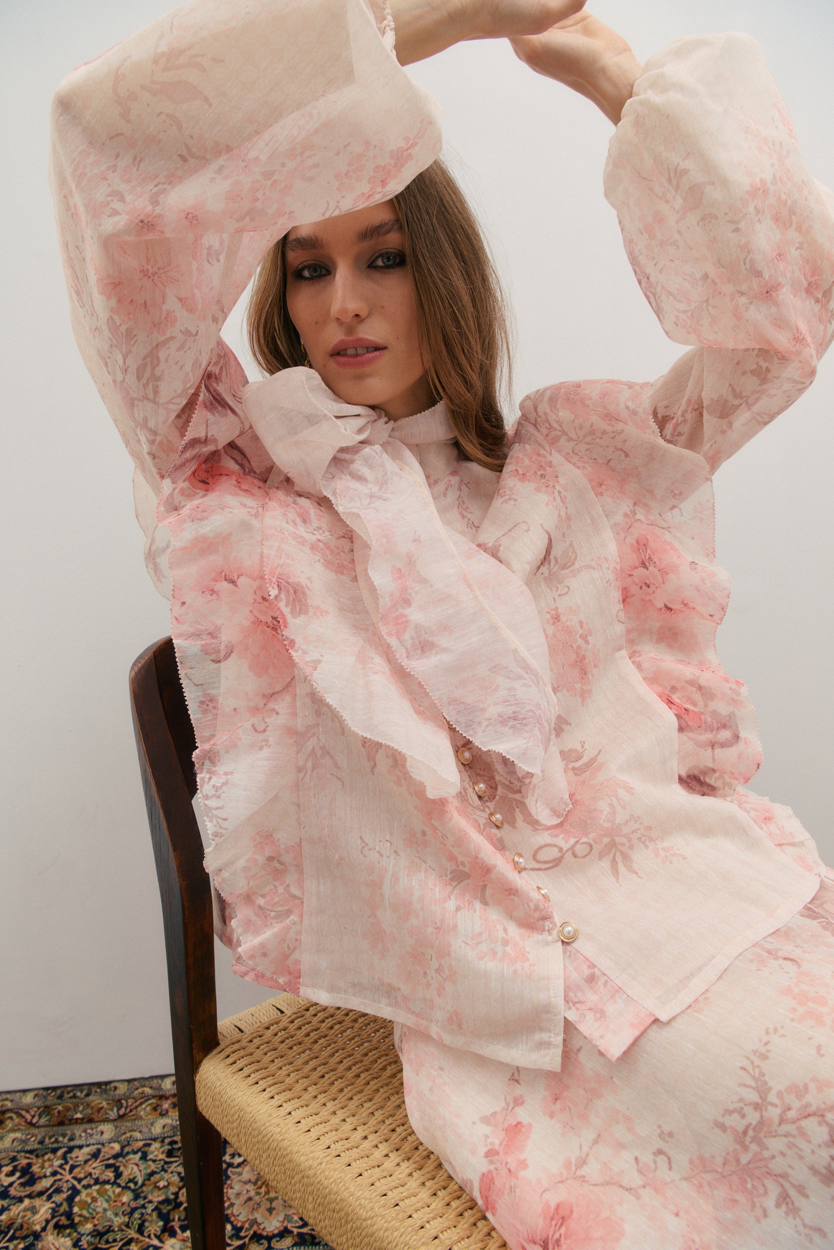 Woman wearing a pink floral outfit sitting on a chair against a plain background