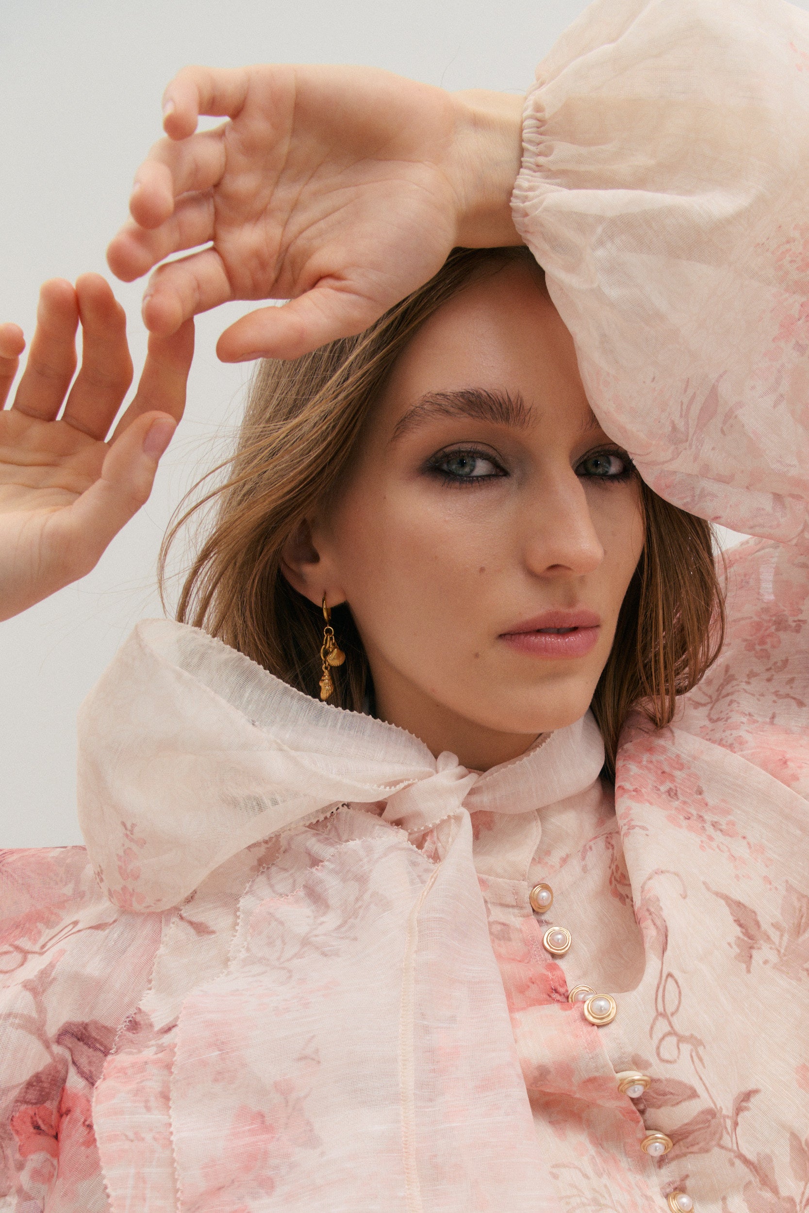 Woman wearing a light pink floral ruffle blouse with long blouson sheer sleeves and elastic cuff, with a large matching scarf tied around her neck against a neutral background
