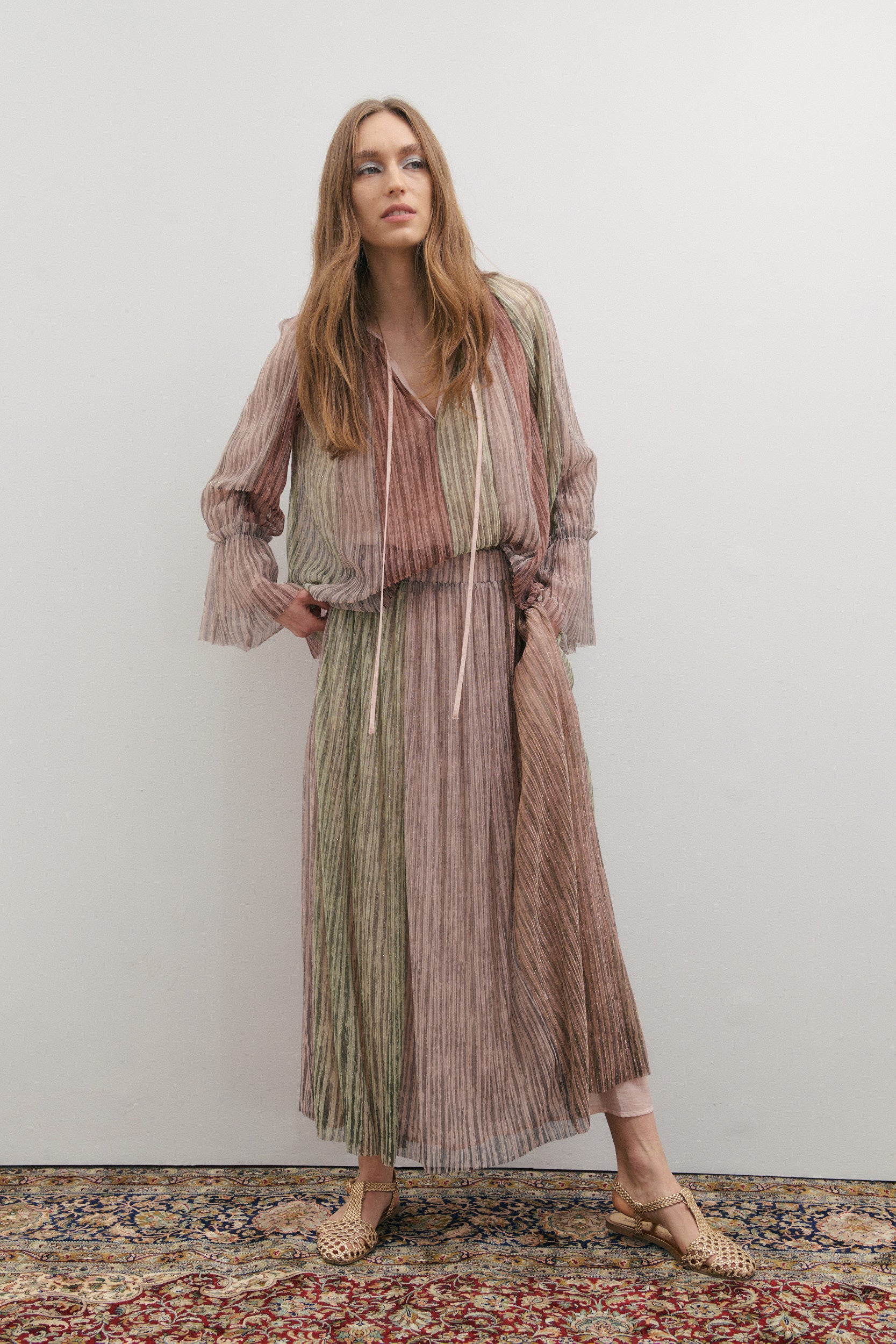 Woman wearing a long, patterned designer outfit standing on a textured rug with a plain background