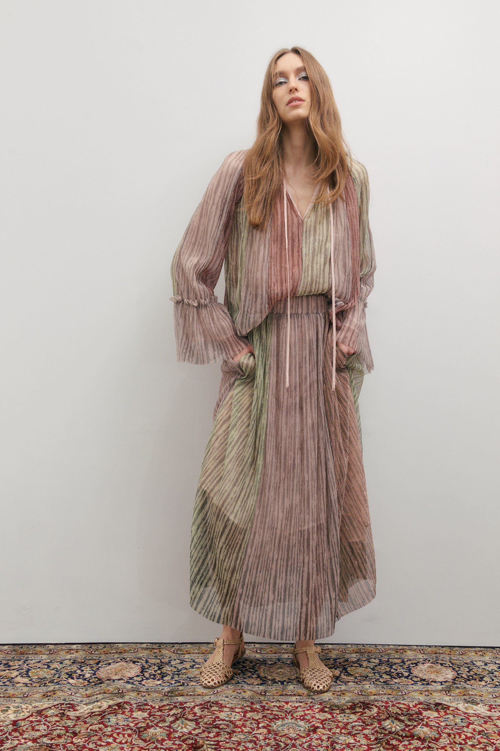 Woman wearing a long, patterned skirt and top standing on a rug with a plain wall background