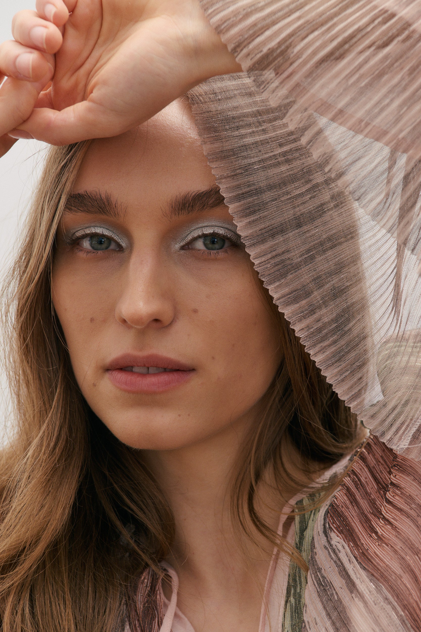 Woman with light makeup and a metallic silver eye, wearing a pink metallic striped top with part of the wide floating sleeve covering her face on a soft background