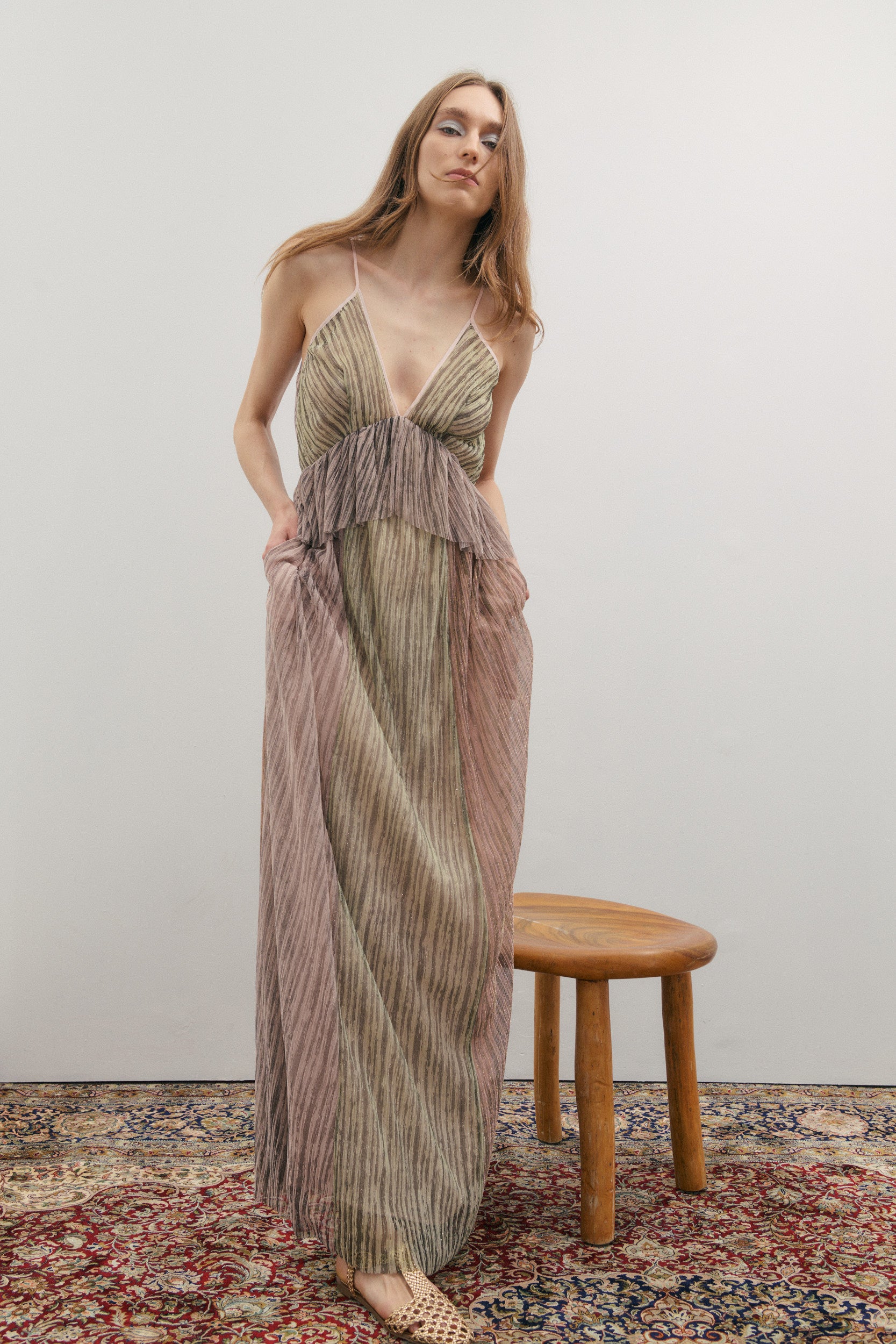 Woman wearing a long, patterned dress standing next to a wooden stool on a patterned rug.