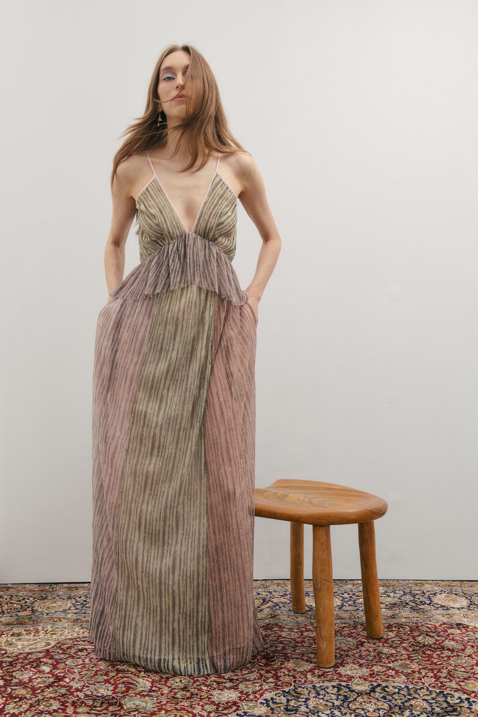 Woman in a long, patterned dress standing next to a wooden stool on a patterned rug.