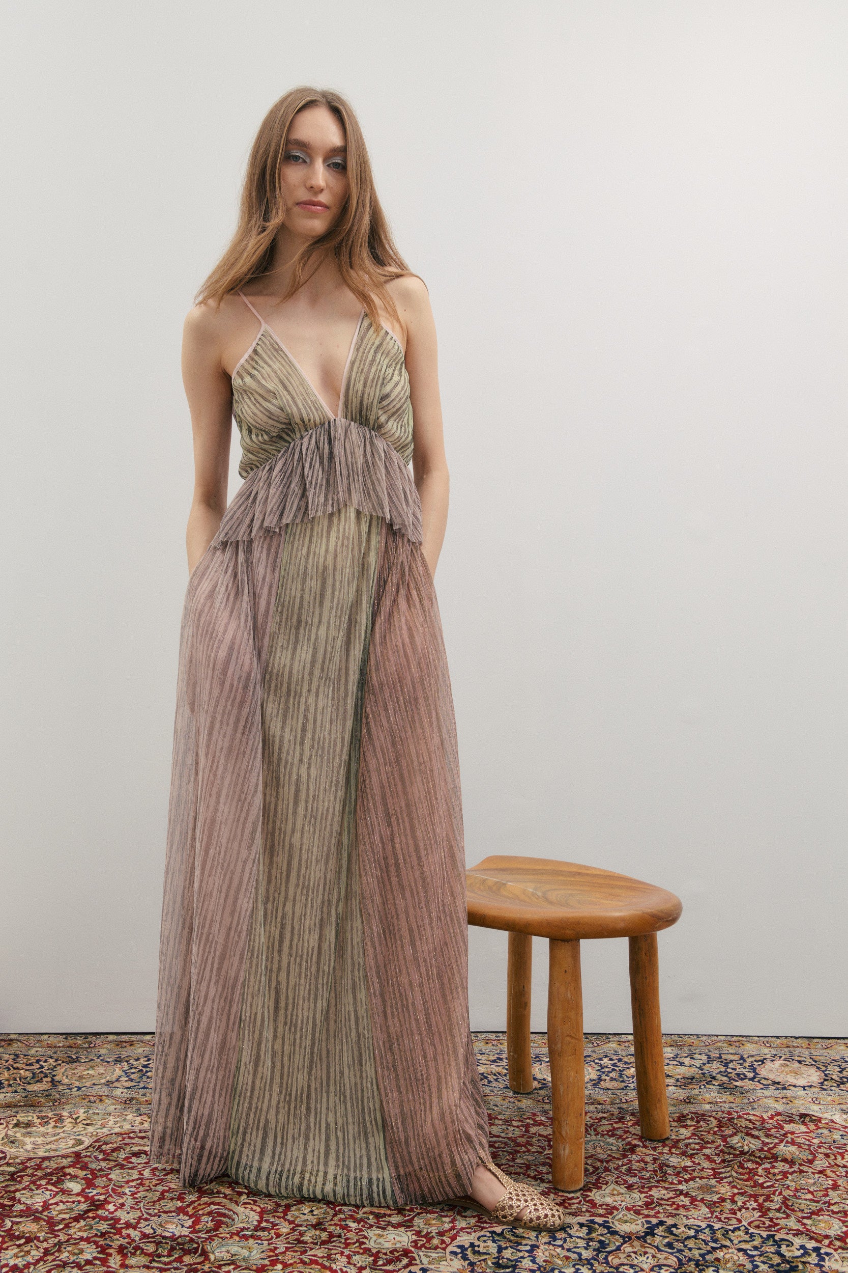 Woman in a long, patterned dress standing next to a wooden stool on a textured rug.