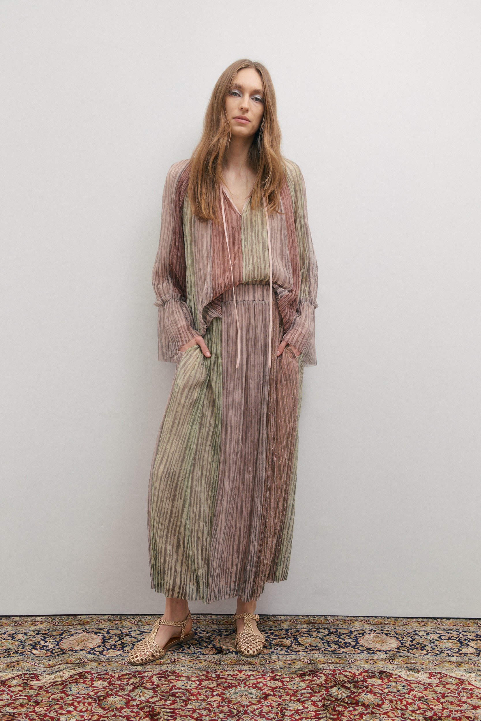 Woman wearing a long, colourful skirt and matching flowing flared sleeves standing against a plain wall.