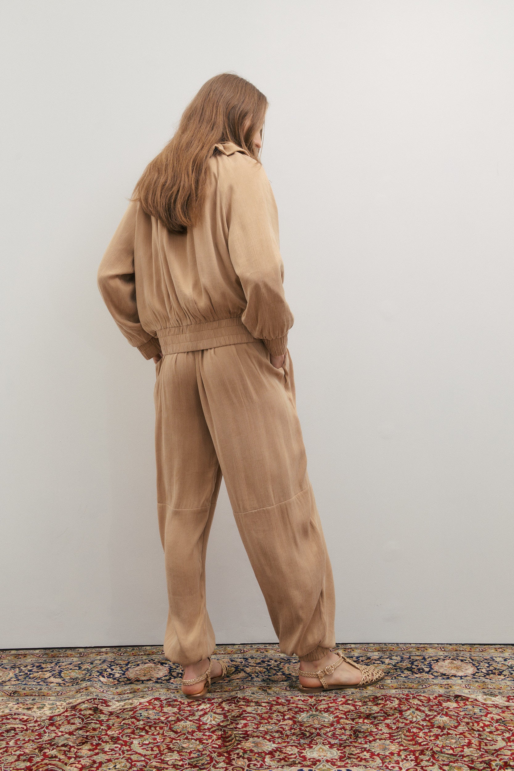Person wearing a beige pant with elastic cuff and matching bomber jacket standing on a patterned rug with a plain wall background