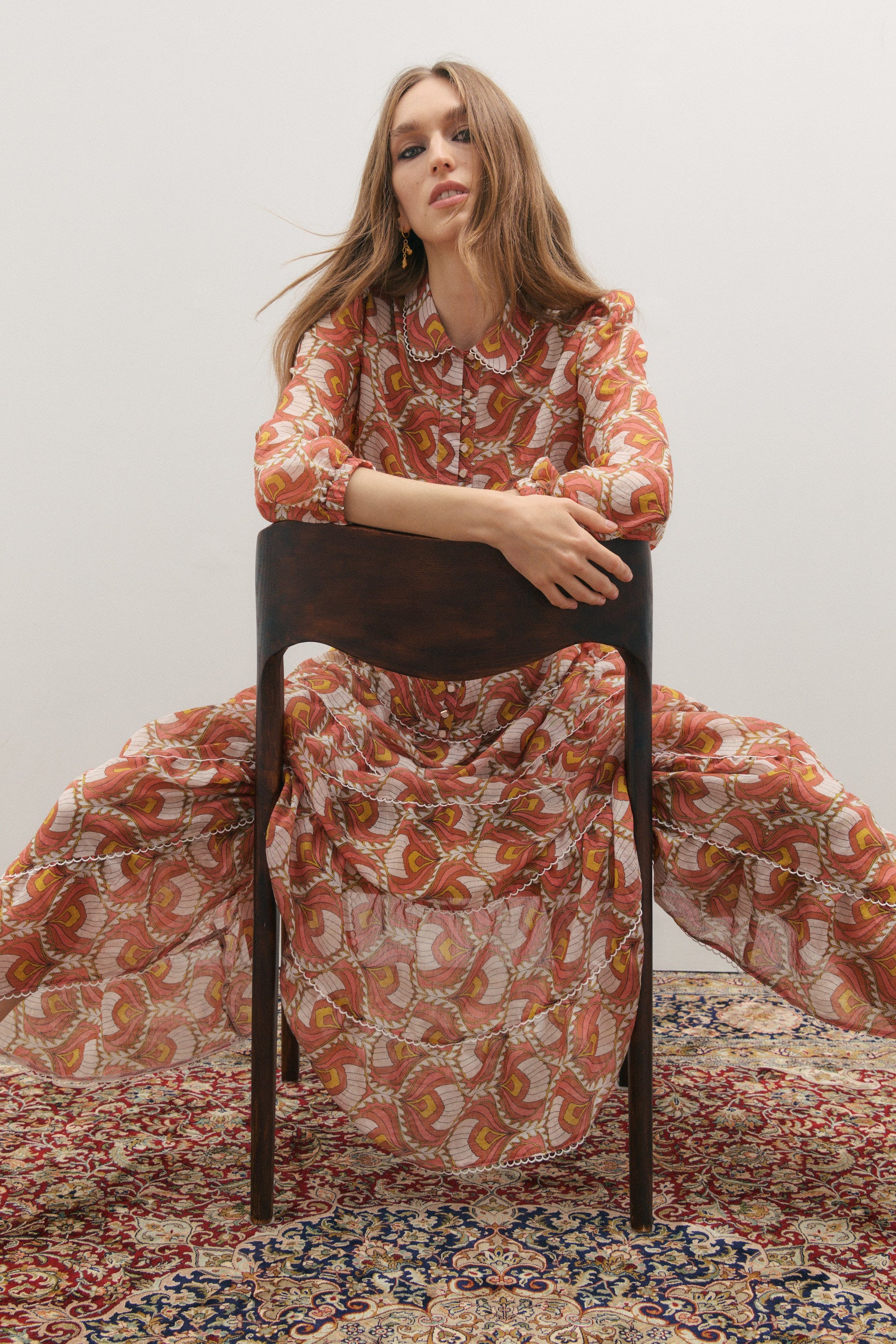 Woman in a patterned dress sitting on a chair against a plain background