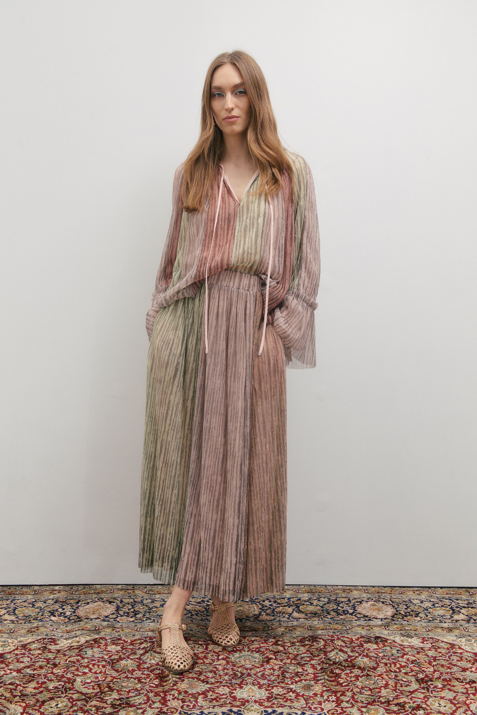 Woman wearing a long, patterned skirt and V-neck blouse standing on a decorative rug with a plain background