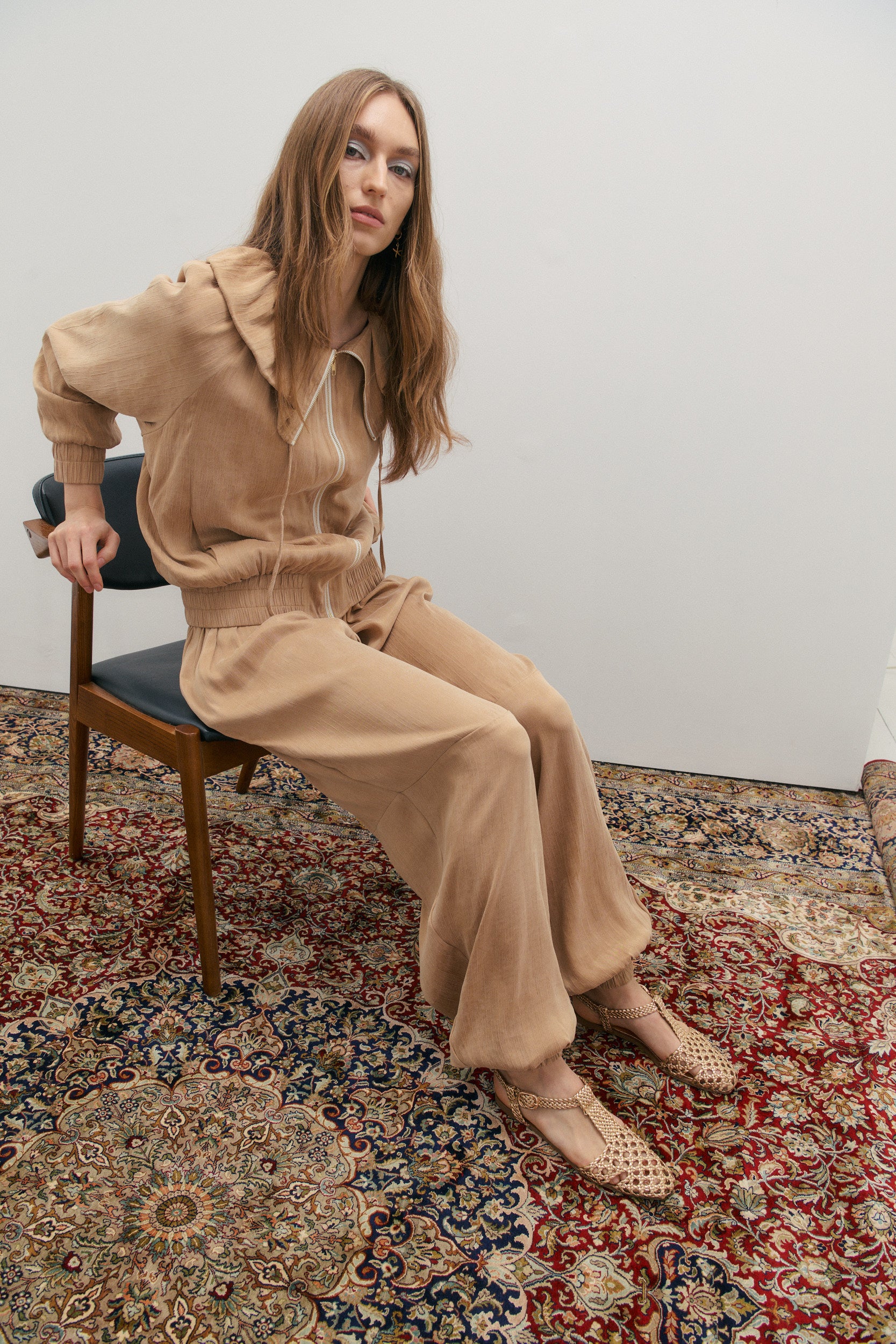 Woman in a beige outfit sitting on a chair with a patterned rug underneath.