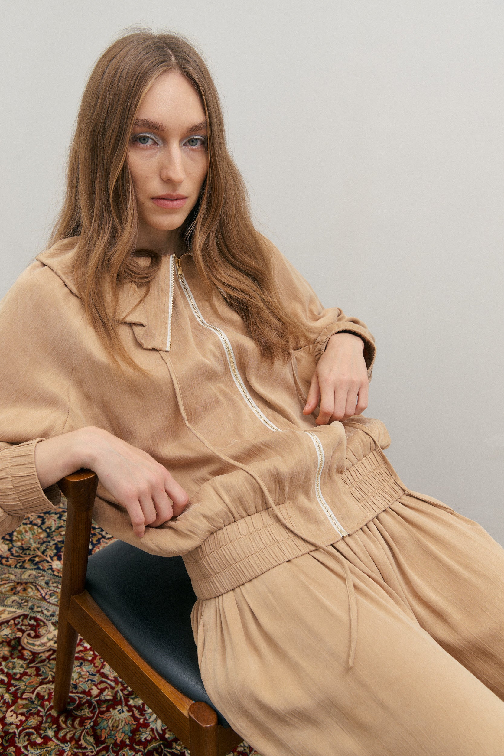 Woman in beige outfit sitting on a chair against a plain background