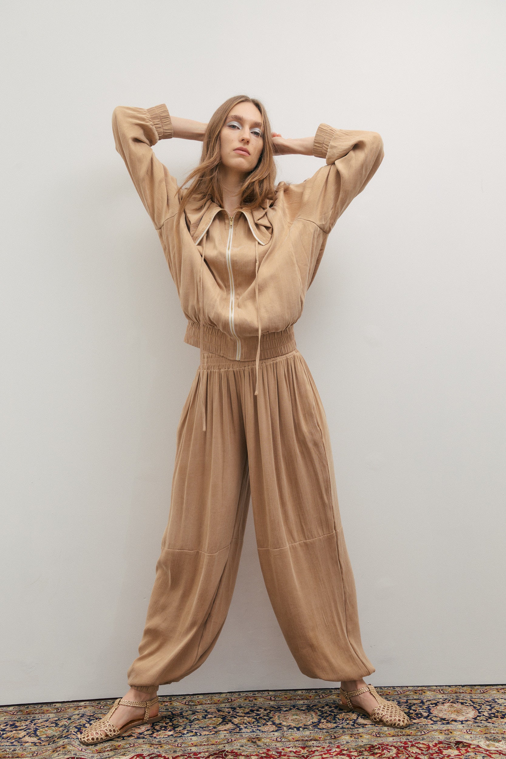 Woman wearing a beige outfit standing against a plain wall.