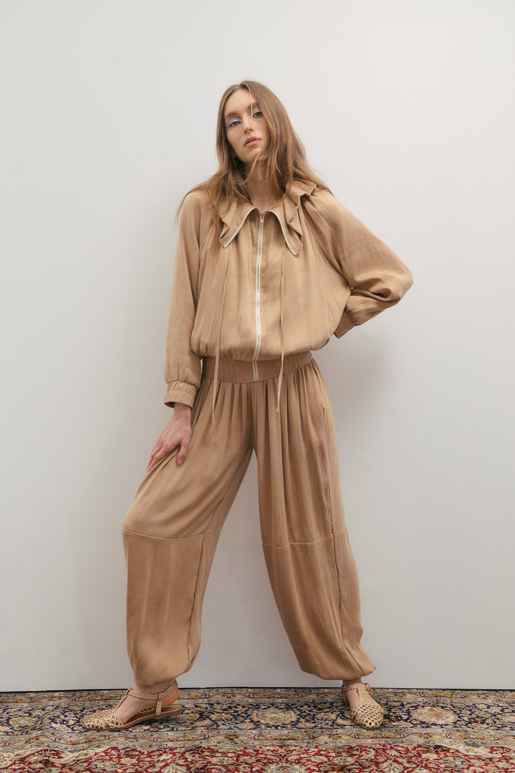 Woman wearing a beige outfit standing against a white wall.