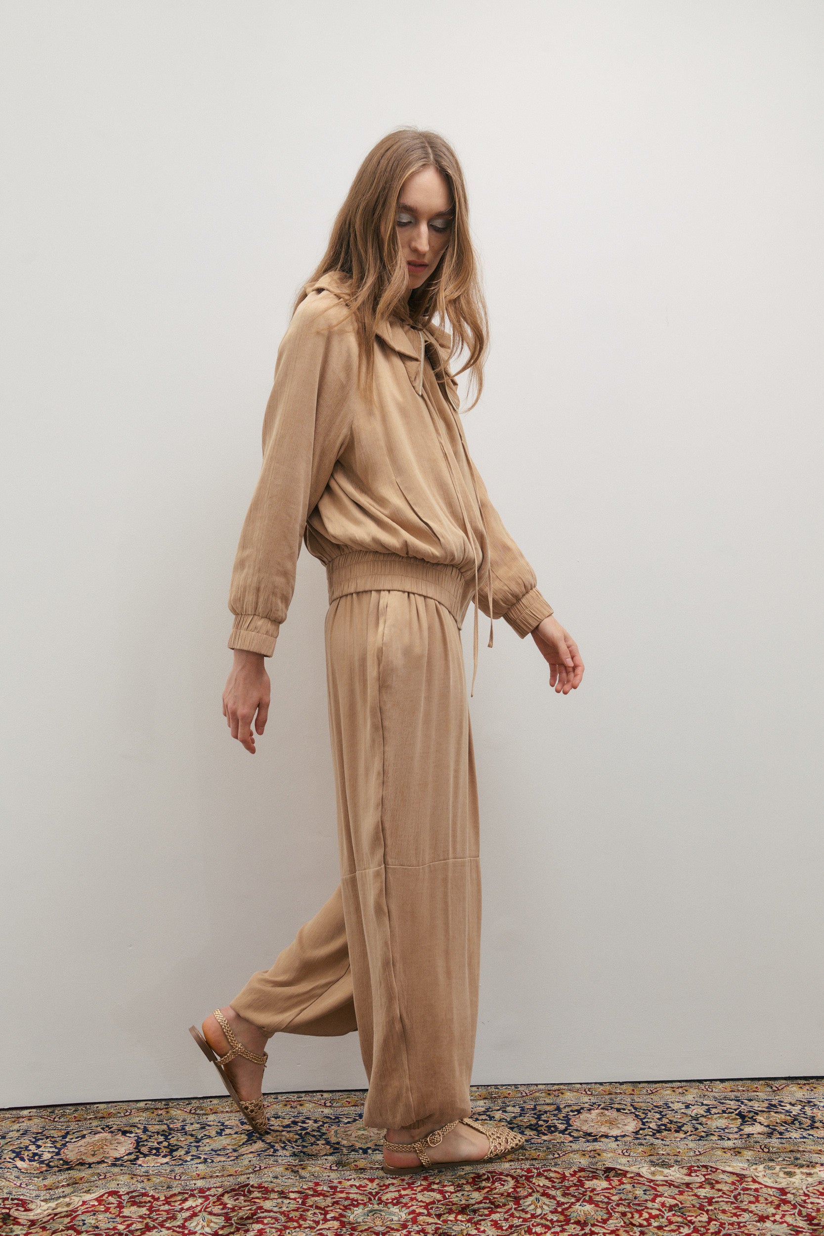 Woman wearing a beige outfit walking on a patterned rug against a plain wall.