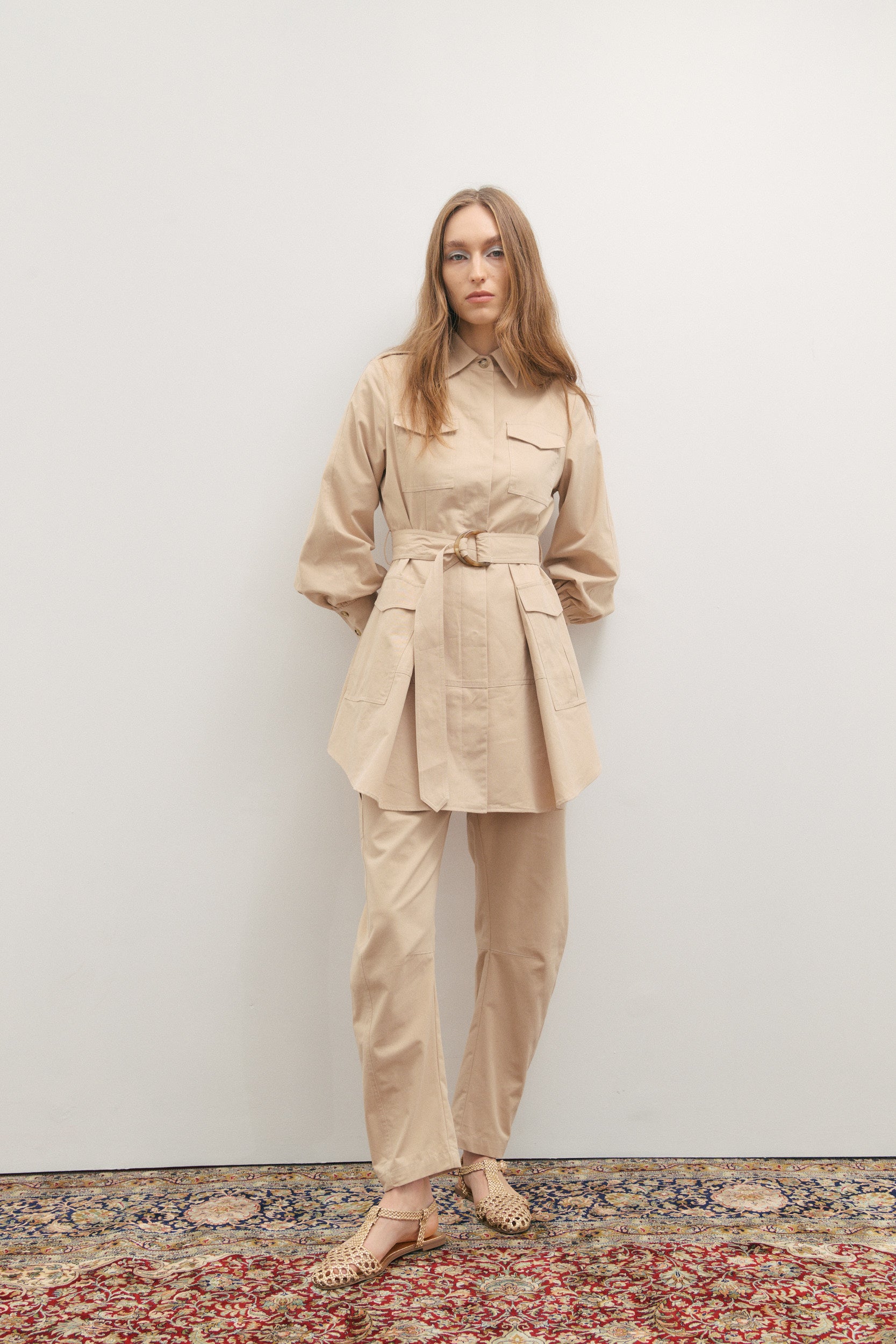 Woman wearing a beige trench coat and pants standing on a patterned rug with a plain wall background