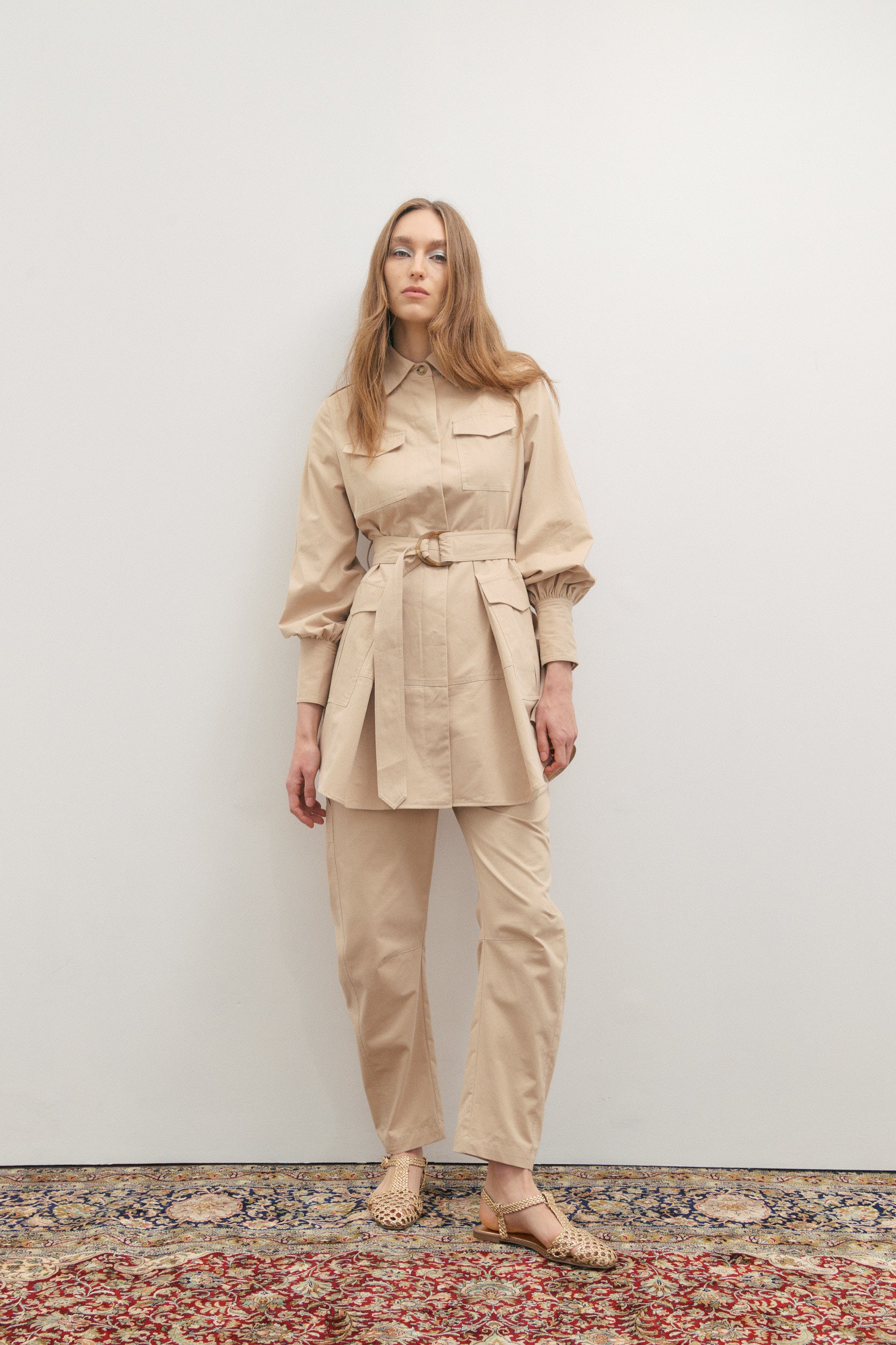 Woman wearing a beige trench coat and pants standing against a white wall.