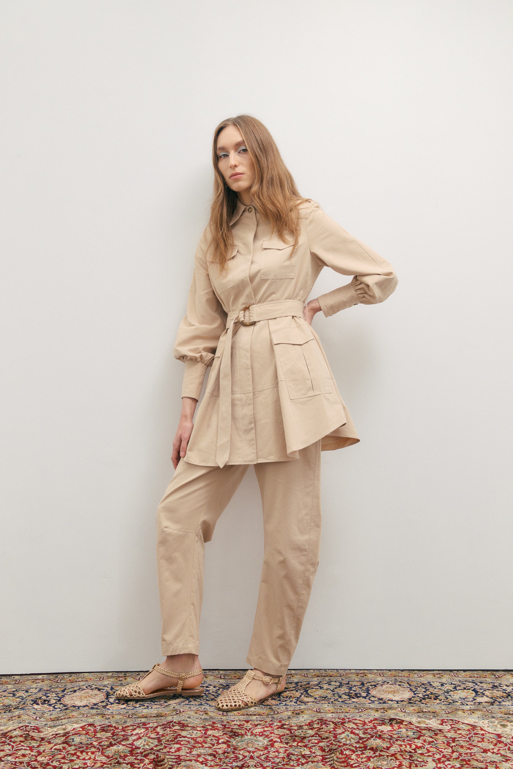 Woman wearing a beige trench coat and pants standing against a plain wall.