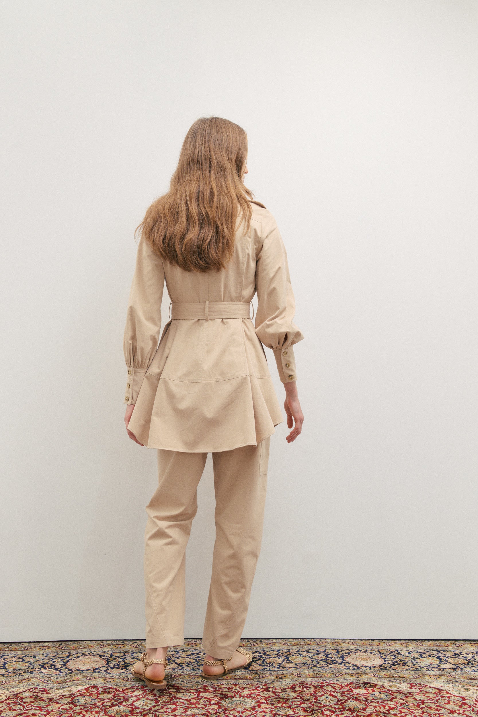 Woman wearing a beige trench coat and pants standing on a patterned rug.