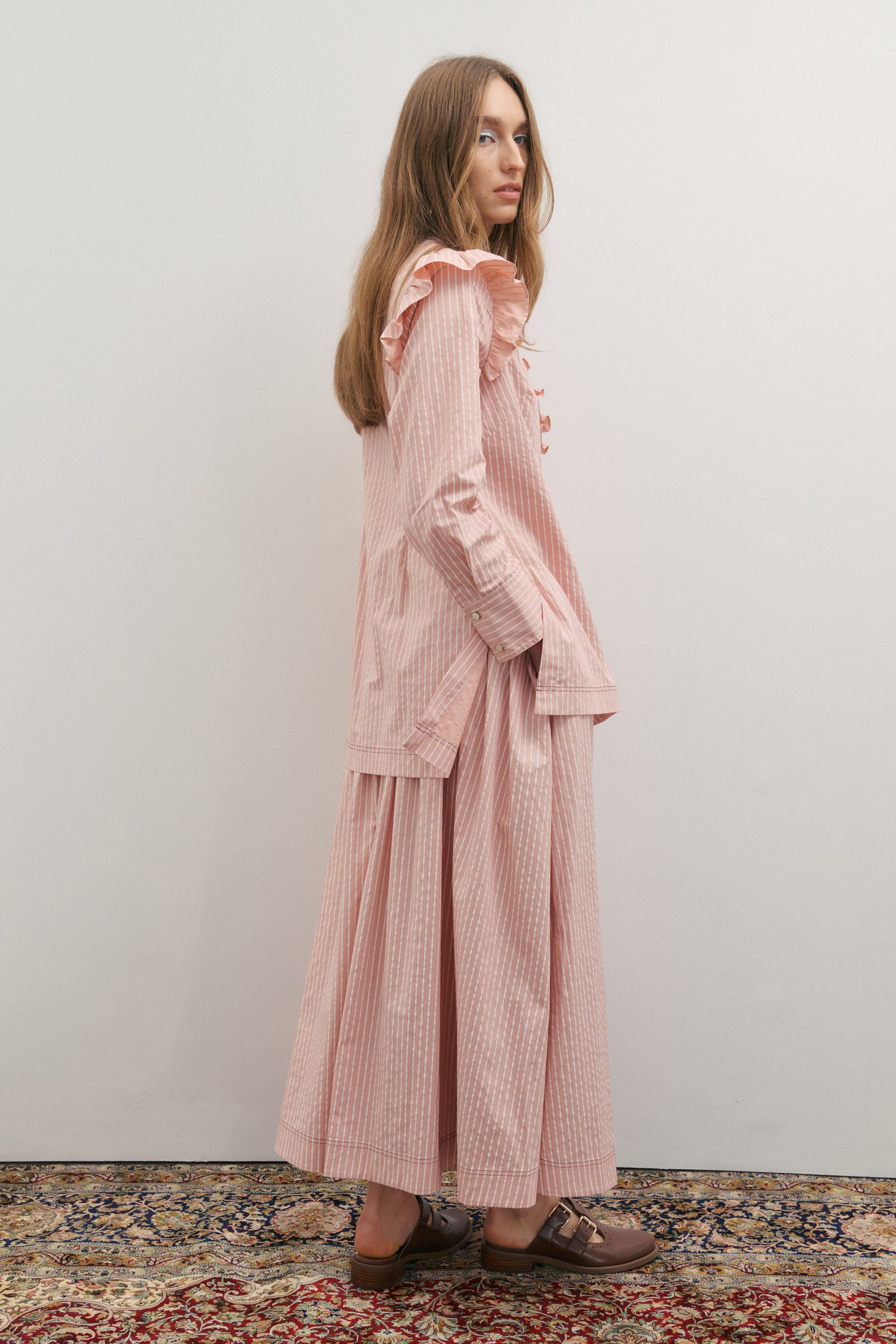 Woman wearing a pink and white striped maxi skirt and shirt outfit standing against a plain wall.