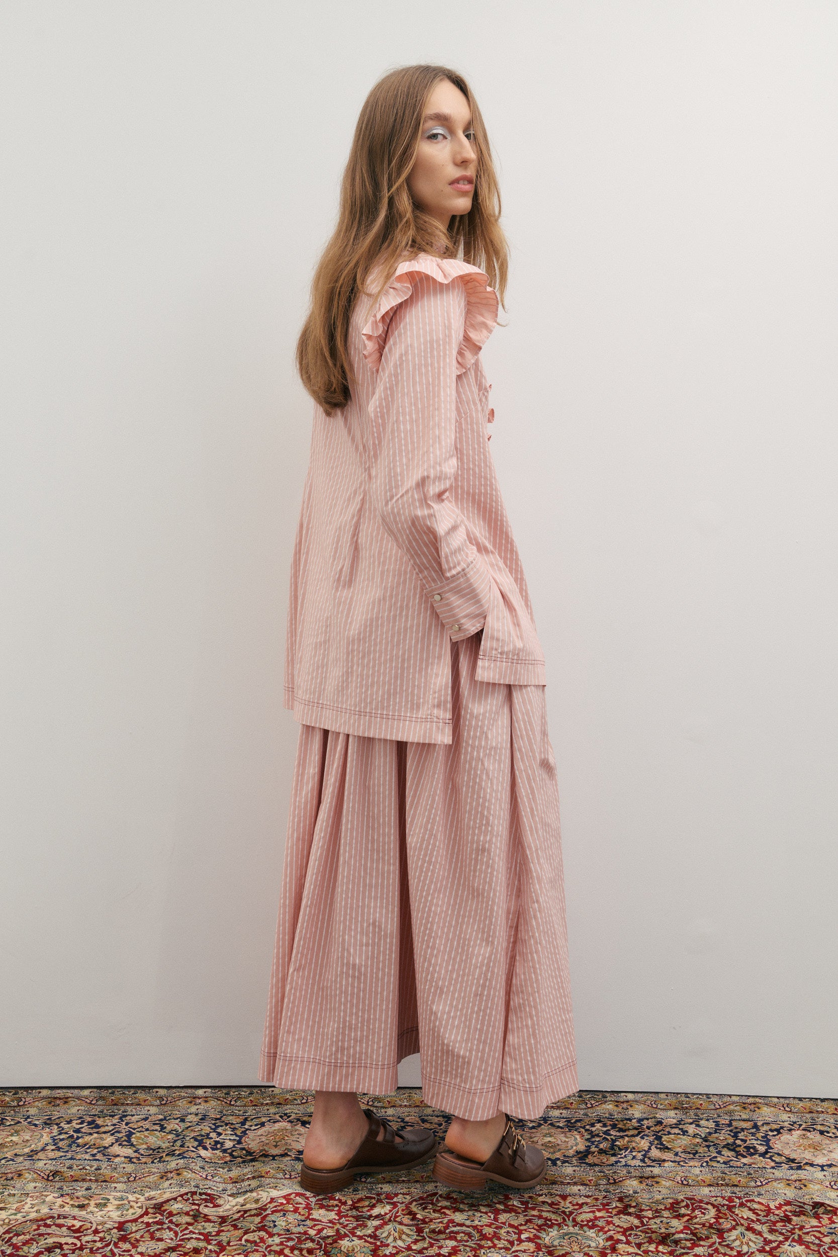 Woman wearing a pink striped outfit standing on a patterned rug with a white wall background