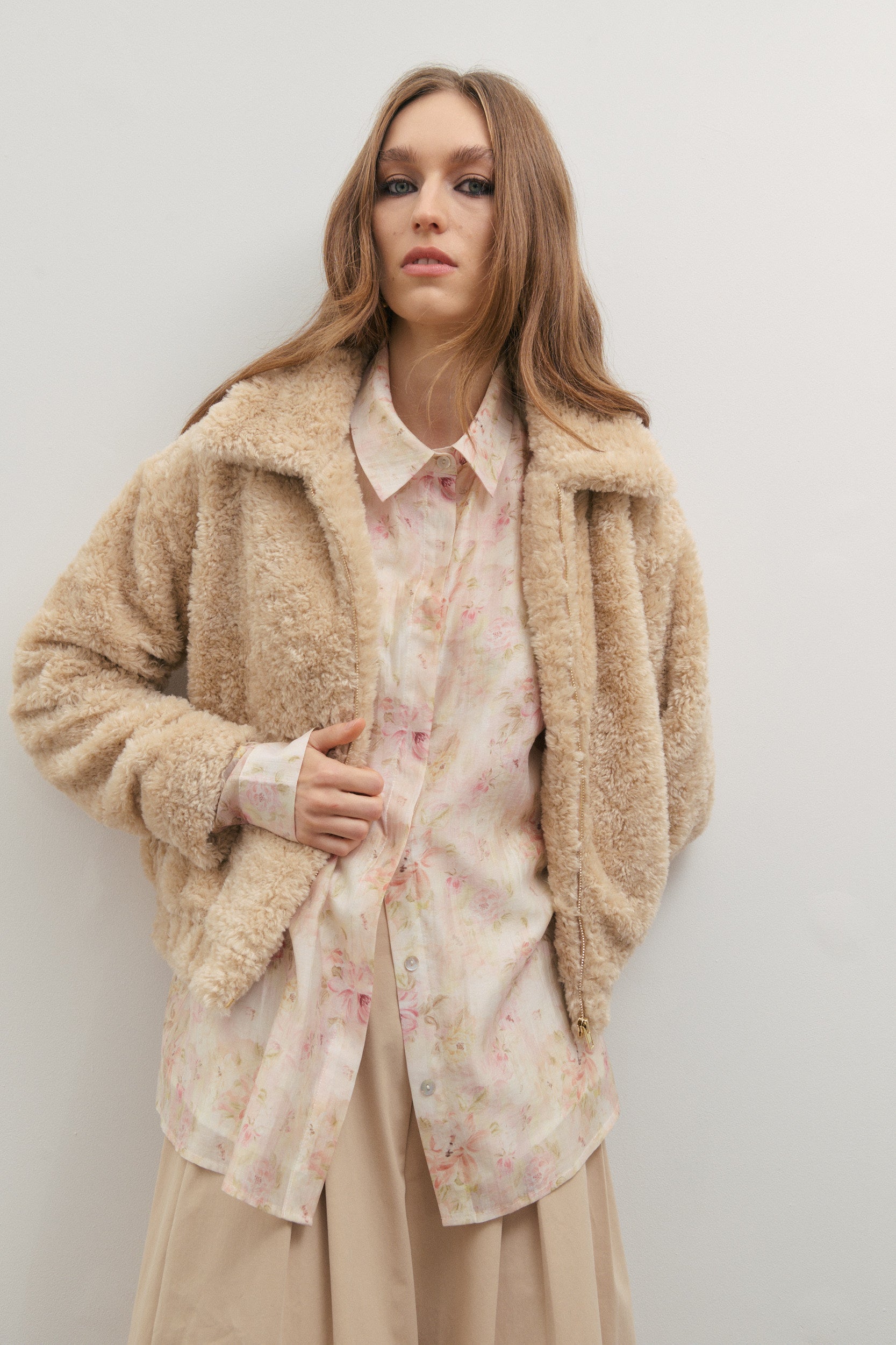Woman wearing a beige teddy coat over a floral shirt and beige pants against a white background