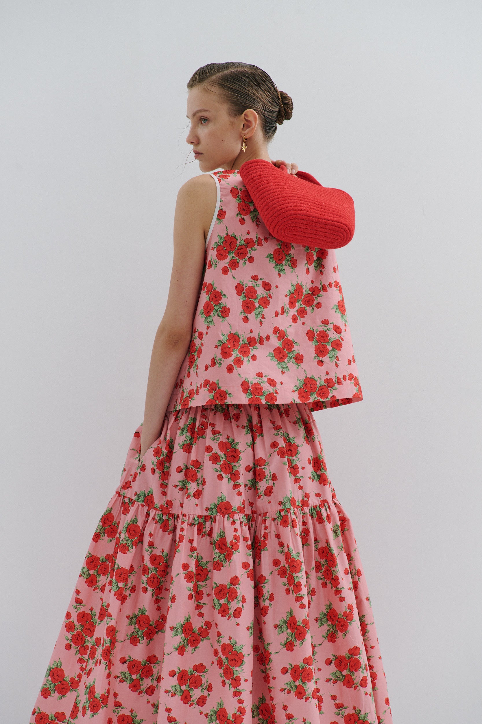 Woman wearing a pink and red floral outfit in a midi skirt and sleeveless top, with a red bag over her shoulder and a hand in a pocket