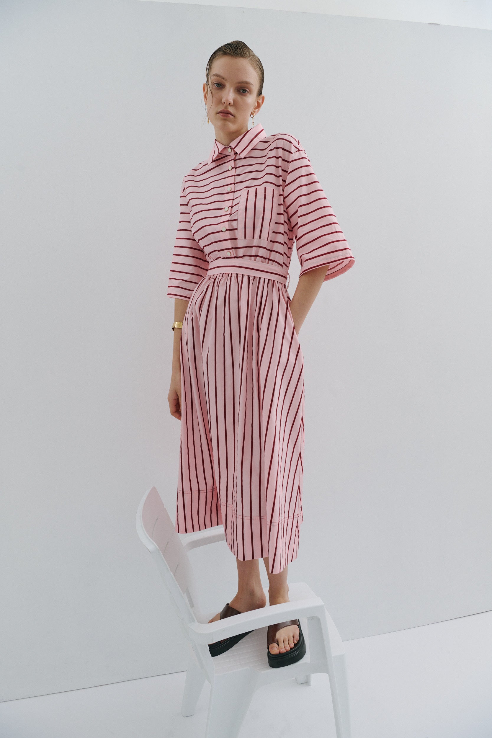 woman wearing a pink and red stripe midi dress with elbow length sleeves, gold pearl buttons, patch pocket, side pockets in a midi length