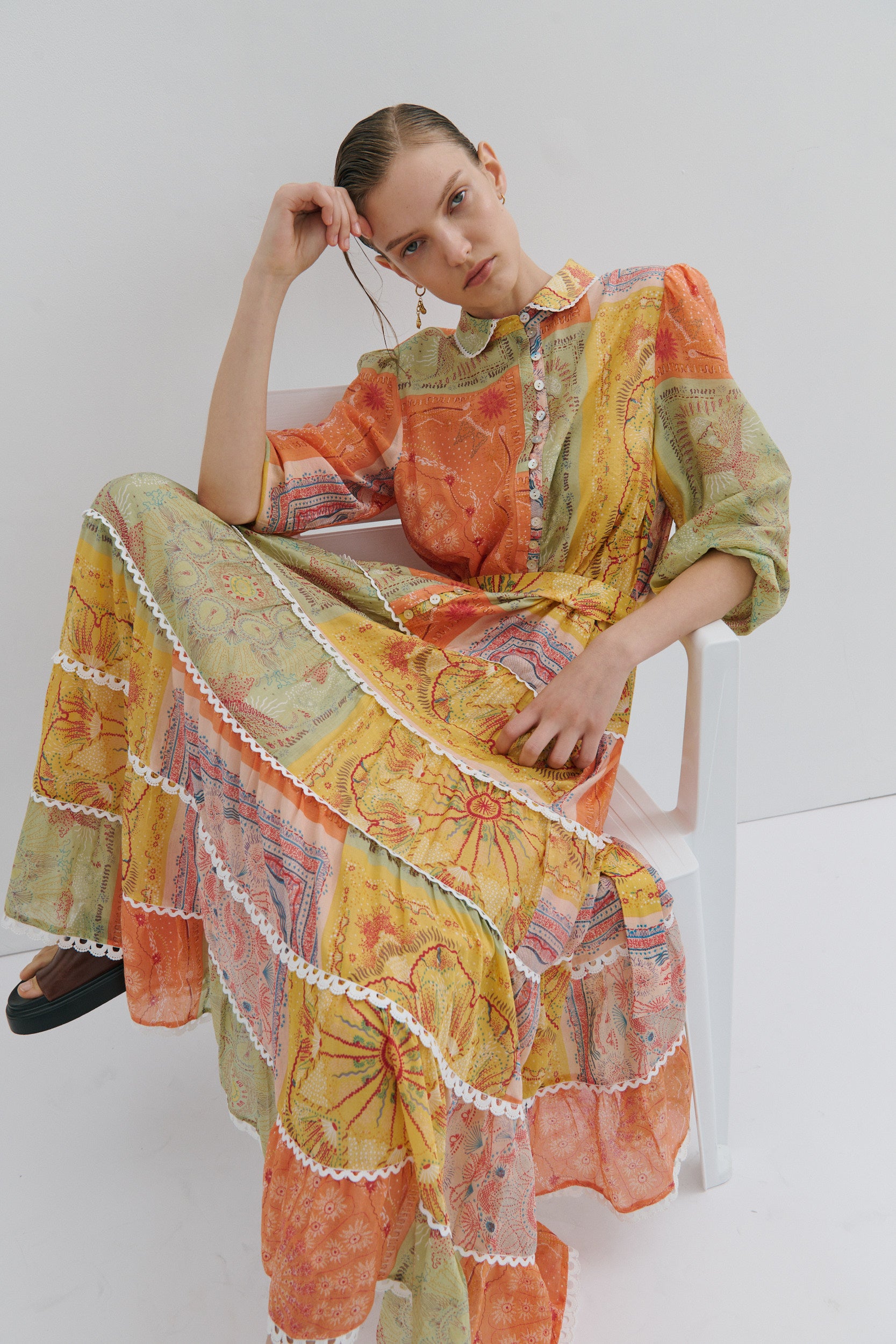Woman wearing a colorful, patterned outfit sitting on a white chair against a white background