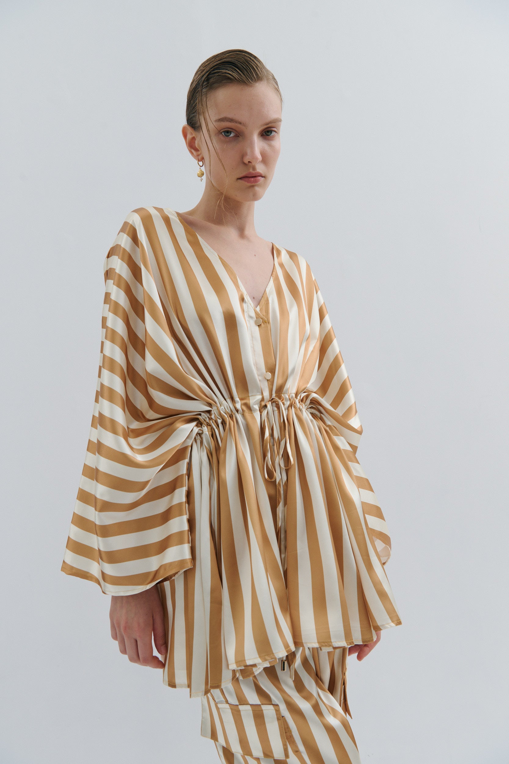 Woman wearing a striped gold and cream kimono style jacket by Australian fashion brand Maevarii, on a plain background