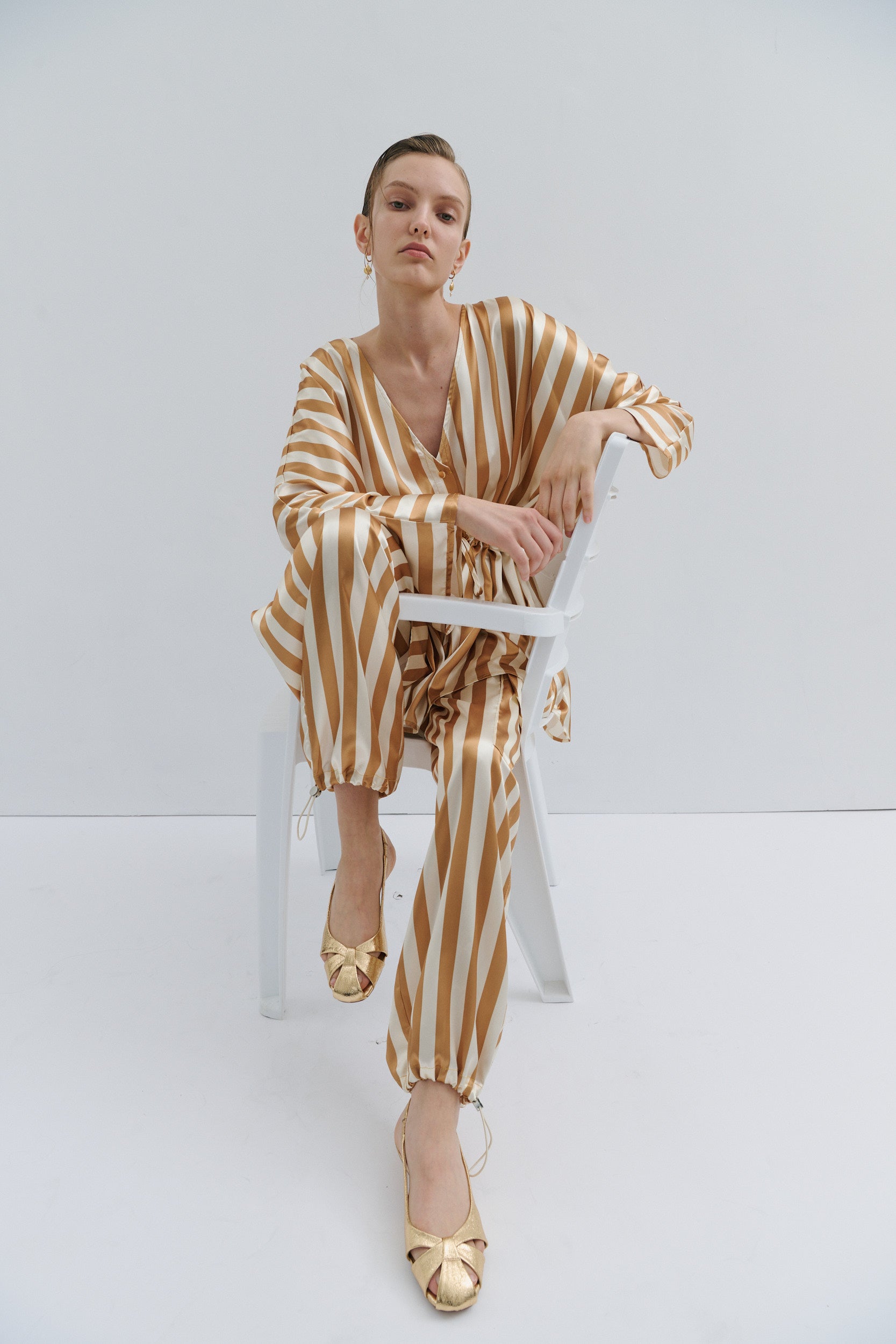 Woman wearing a gold and white striped Maevarii outfit sitting on a white chair against a white background