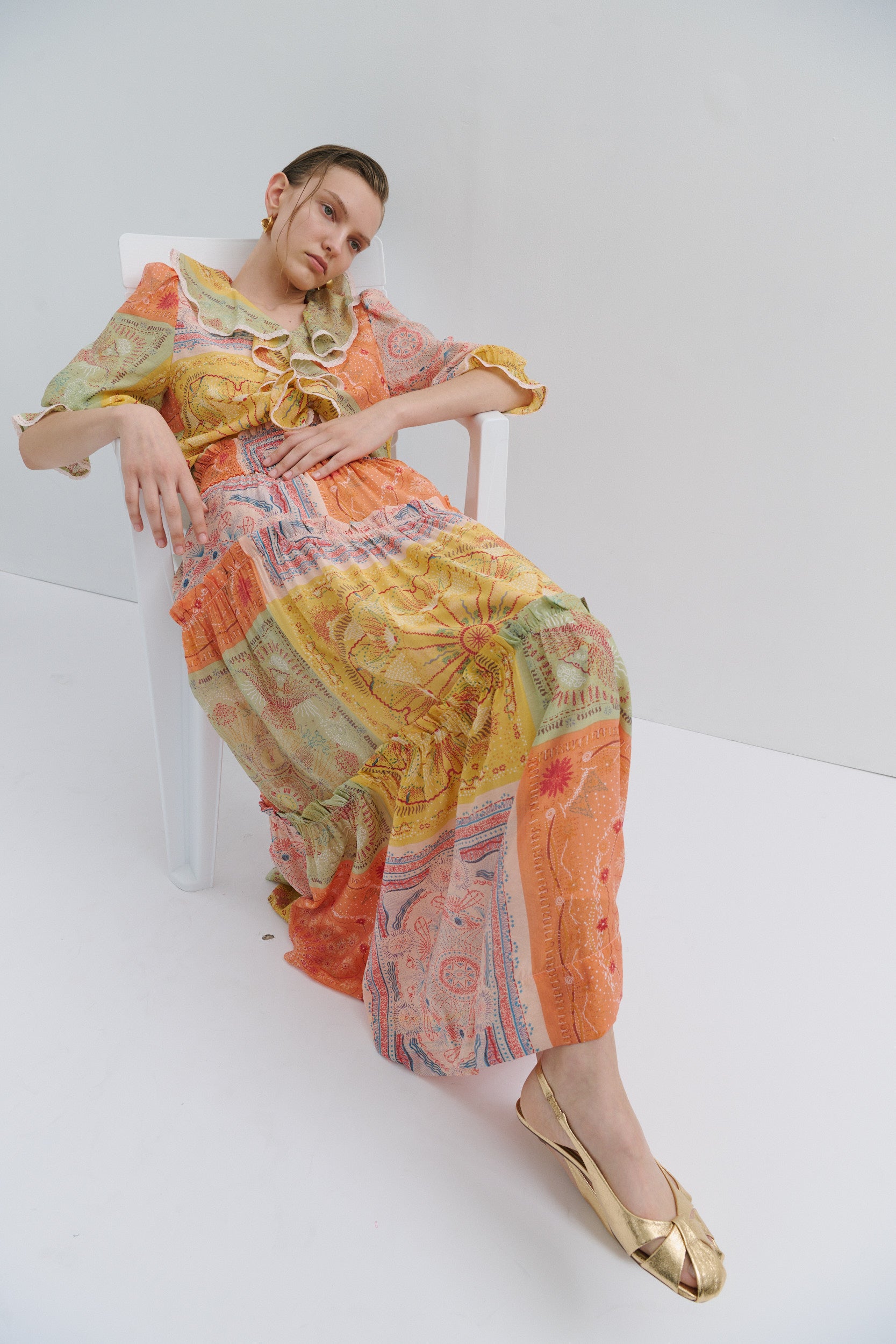 Woman in a colorful skirt and top sitting on a white chair against a white background