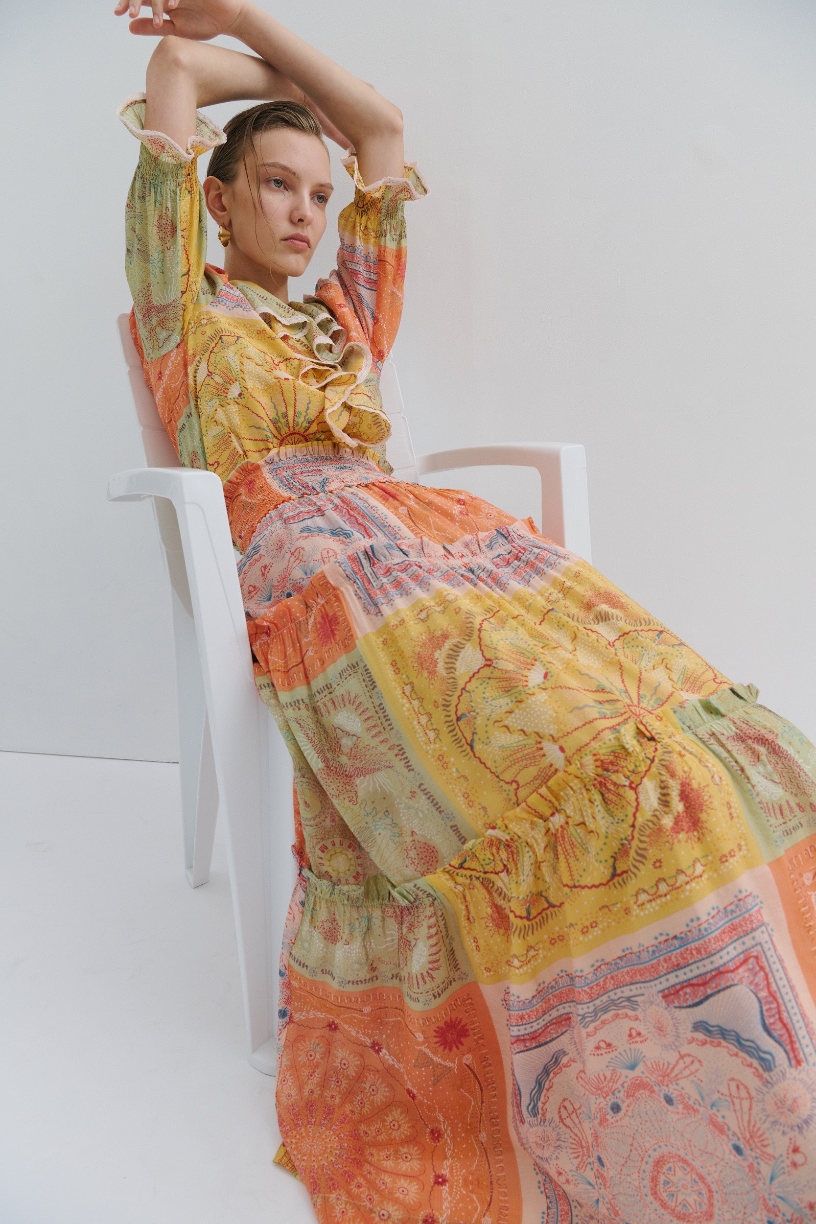 Woman wearing a colorful patterned boho frill shirt and long tiered maxi skirt with smocked waistband sitting on a white chair against a white background