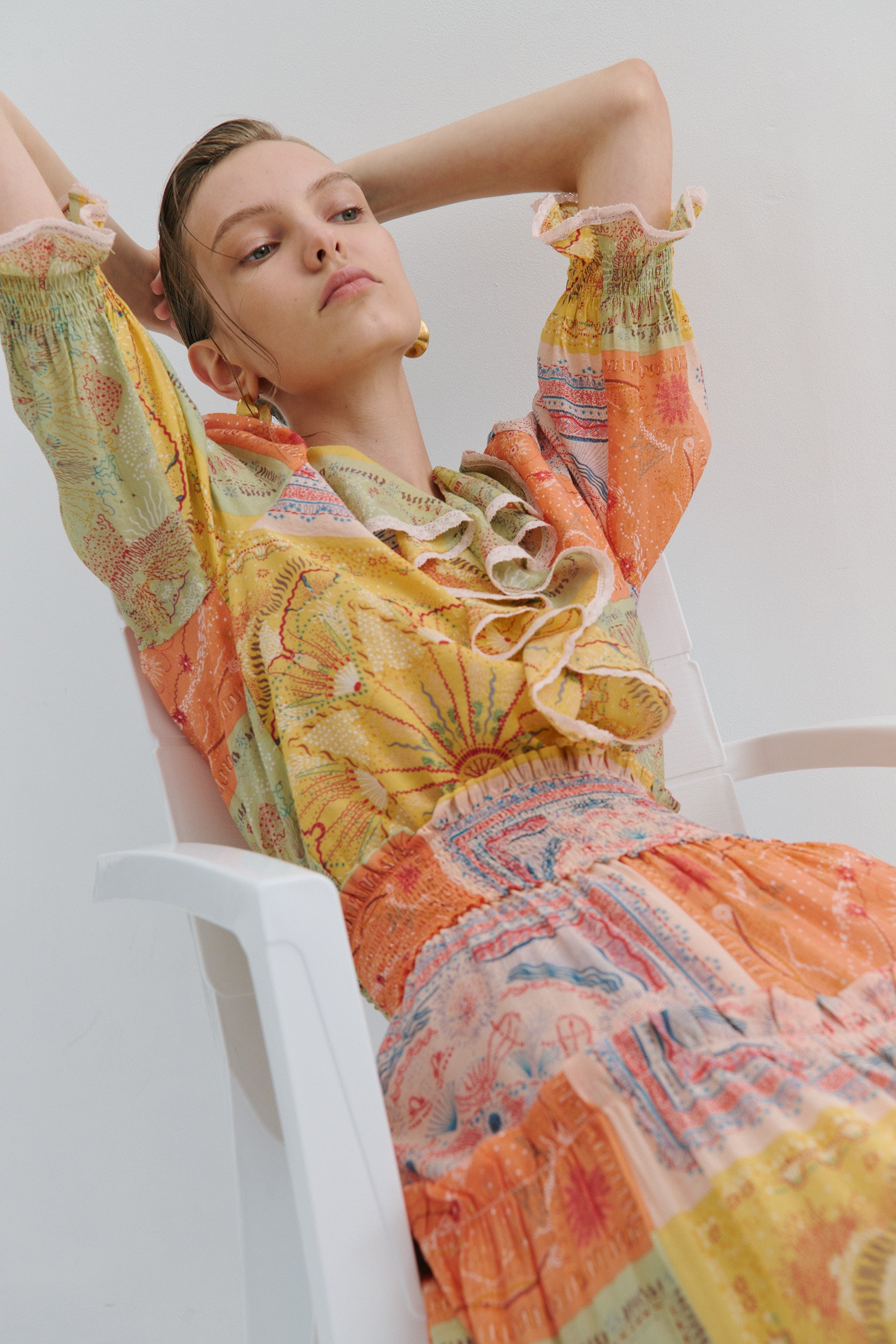 Woman wearing a colorful patterned boho frill blouse by Australian fashion brand Maevarii, sitting on a white chair against a light background