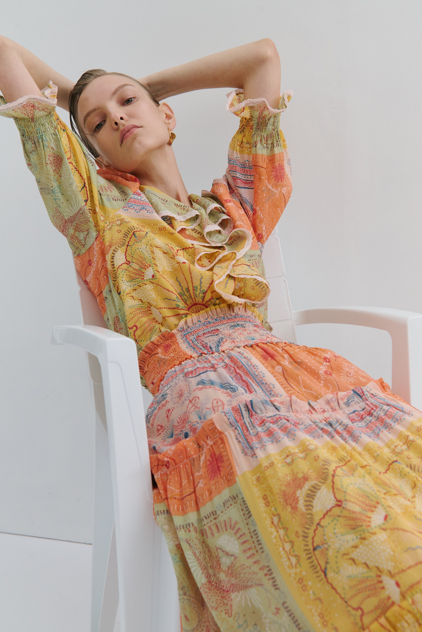 Woman wearing a colourful top and skirt sitting on a white chair against a white background