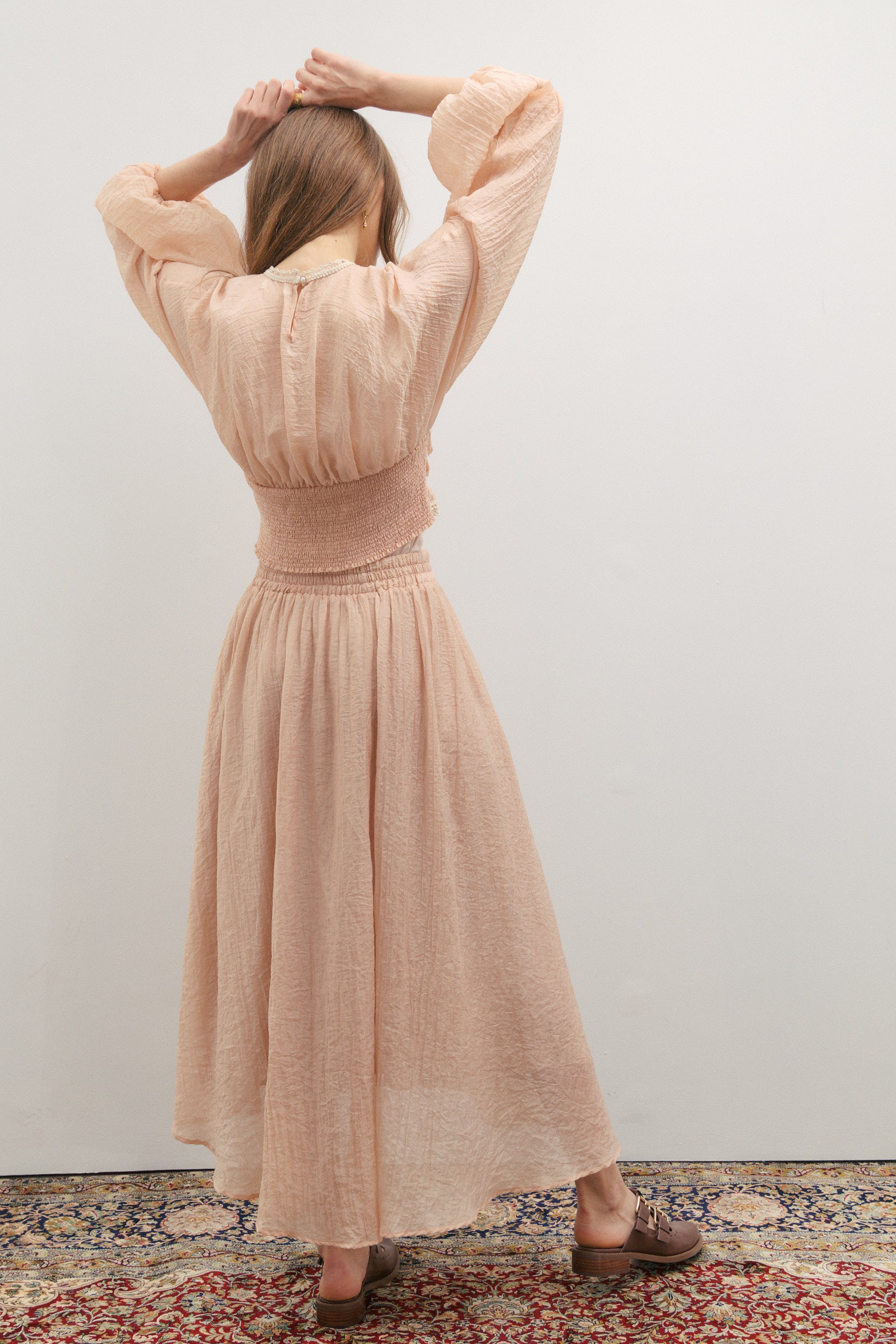 Woman wearing a soft peach pearl colour long skirt and matching top with billowing sleeves with an elastic cuff and a cream embroidery bodice with soft elastic smocking at the back by Australian womenswear brand Maevarii, while standing against a white wall.