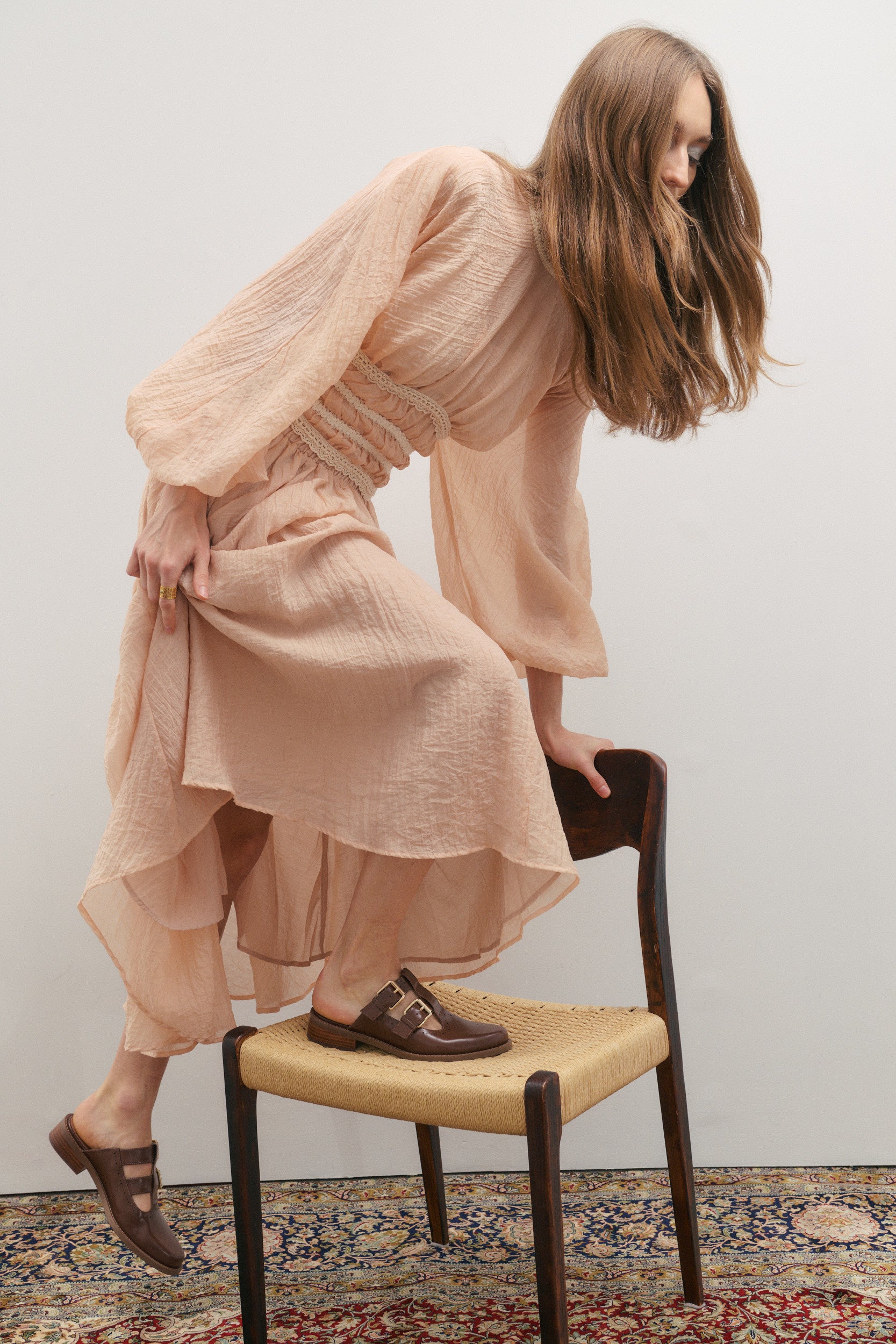 Woman in a flowing long outfit by womenswear brand Maevarii sitting on a chair against a plain background